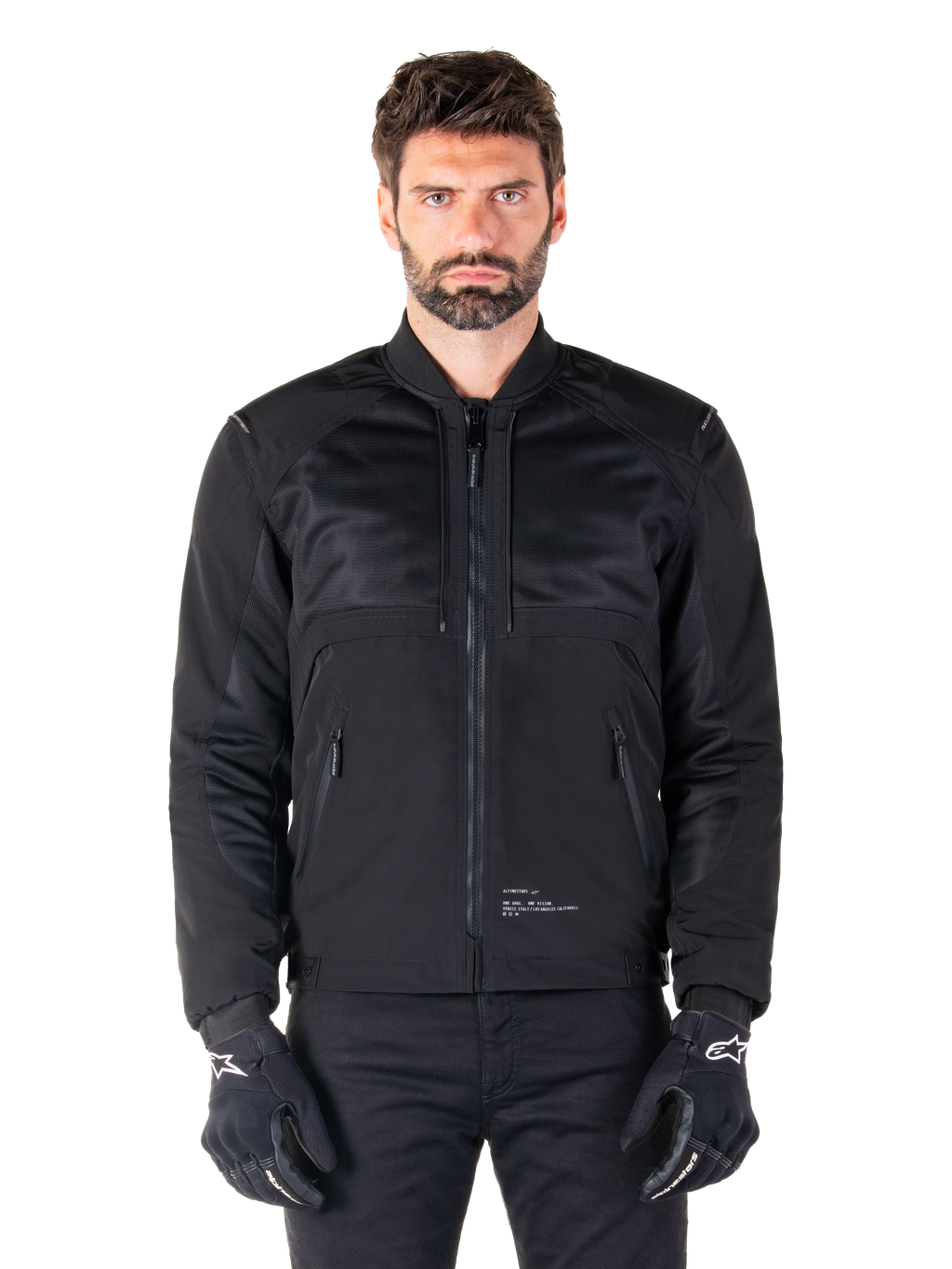 Alpinestars Mootant Waterproof Parka, zwarte motorjas, vooraanzicht op mannelijk model, met modulaire textielconstructie met mesh-panelen voor ventilatie, geribbelde kraag en zakken met ritssluiting, gedragen met zwarte handschoenen met witte Alpinestars-branding