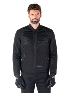Alpinestars Mootant Waterproof Parka, zwarte motorjas, vooraanzicht op mannelijk model, met modulaire textielconstructie met mesh-panelen voor ventilatie, geribbelde kraag en zakken met ritssluiting, gedragen met zwarte handschoenen met witte Alpinestars-branding