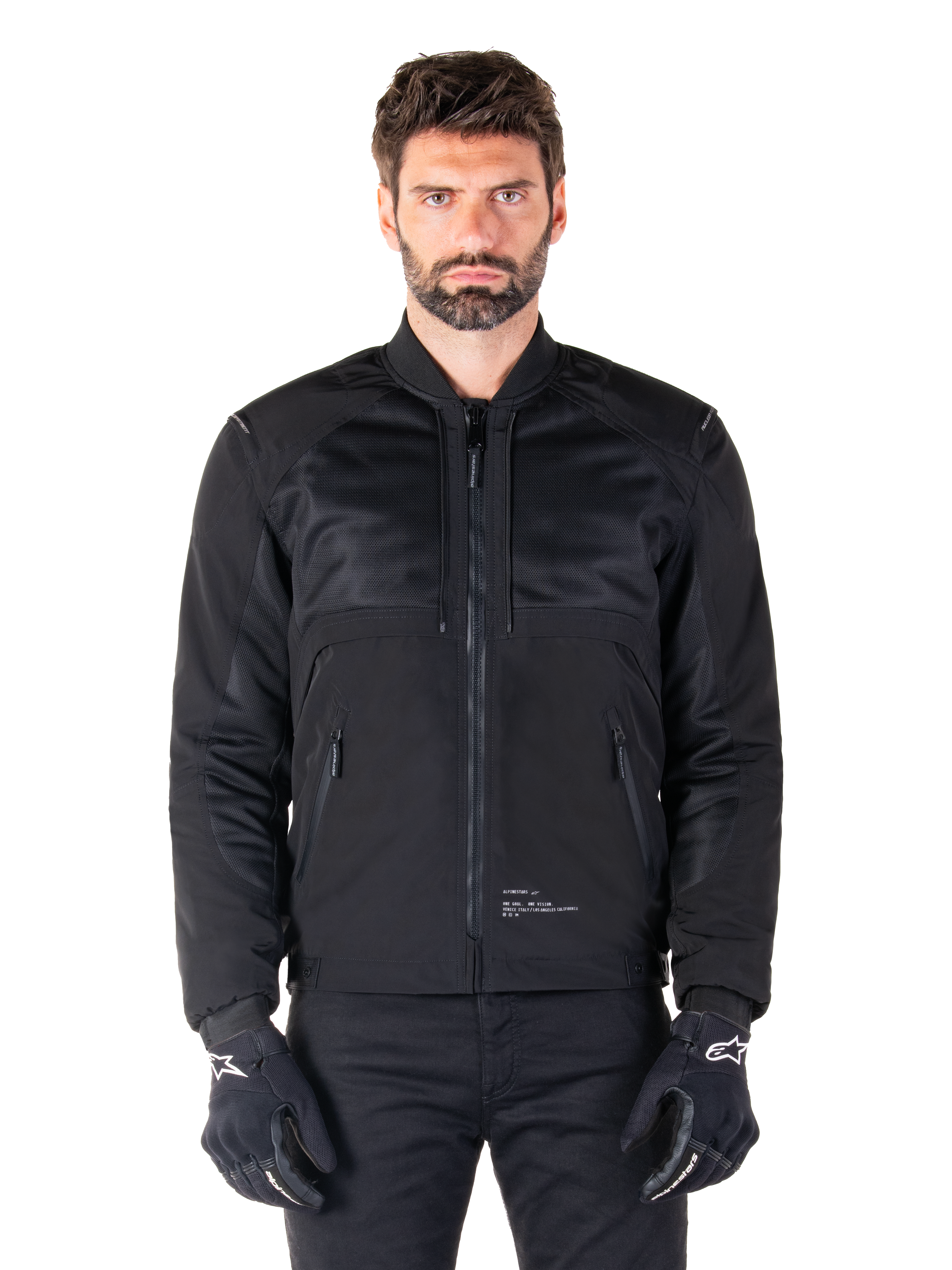 Alpinestars Mootant Waterproof Parka, zwarte motorjas, vooraanzicht op mannelijk model, met modulaire textielconstructie met mesh-panelen voor ventilatie, geribbelde kraag en zakken met ritssluiting, gedragen met zwarte handschoenen met witte Alpinestars-branding