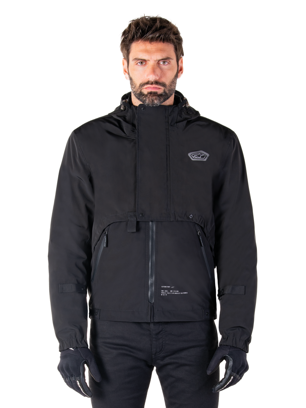 Alpinestars Mootant Waterproof Parka, zwarte motorjas met capuchon en een modulair technisch ontwerp, gedragen door een mannelijk model met zwarte handschoenen, voorzien van een unieke borstflap en zijzakken met rits, urban en touring rijstijl