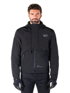 Alpinestars Mootant Waterproof Parka, zwarte motorjas met capuchon en een modulair technisch ontwerp, gedragen door een mannelijk model met zwarte handschoenen, voorzien van een unieke borstflap en zijzakken met rits, urban en touring rijstijl