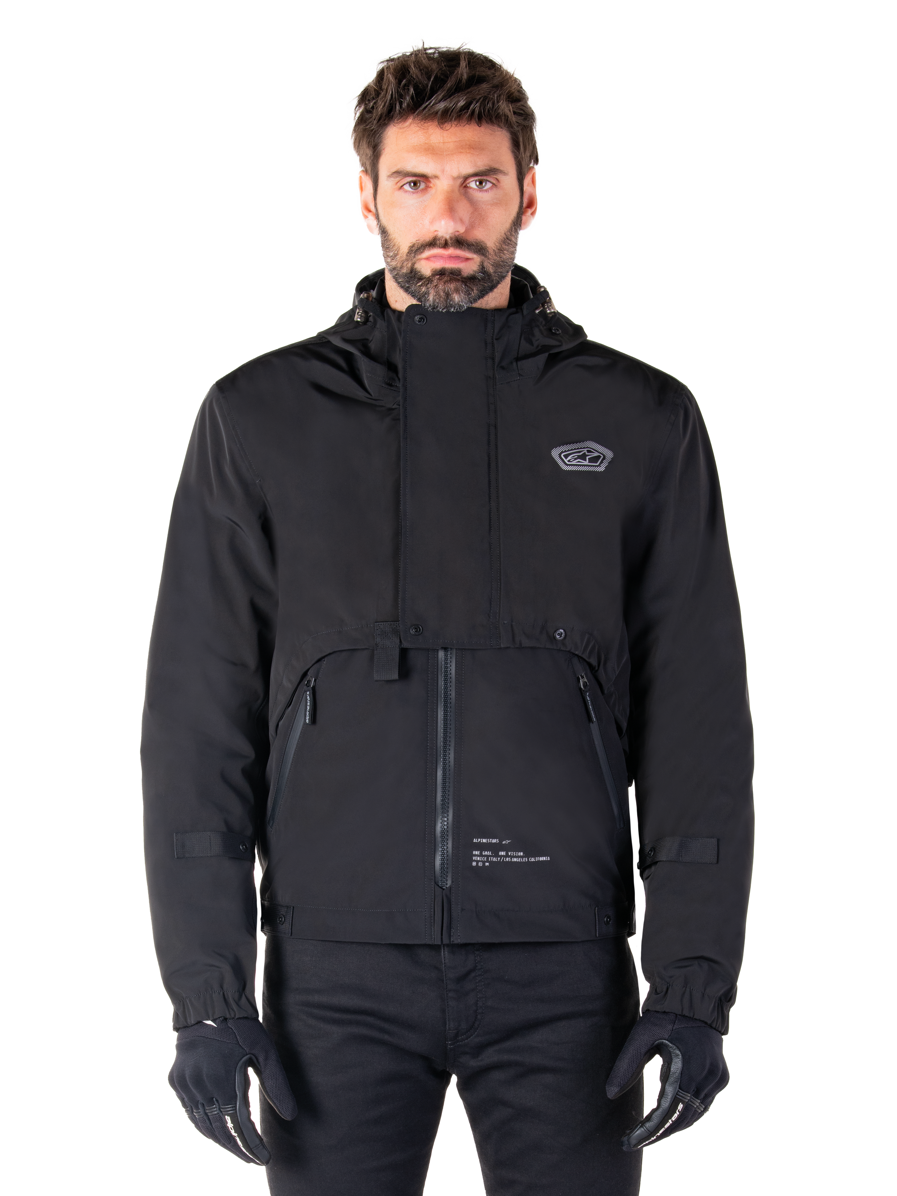 Alpinestars Mootant Waterproof Parka, zwarte motorjas met capuchon en een modulair technisch ontwerp, gedragen door een mannelijk model met zwarte handschoenen, voorzien van een unieke borstflap en zijzakken met rits, urban en touring rijstijl