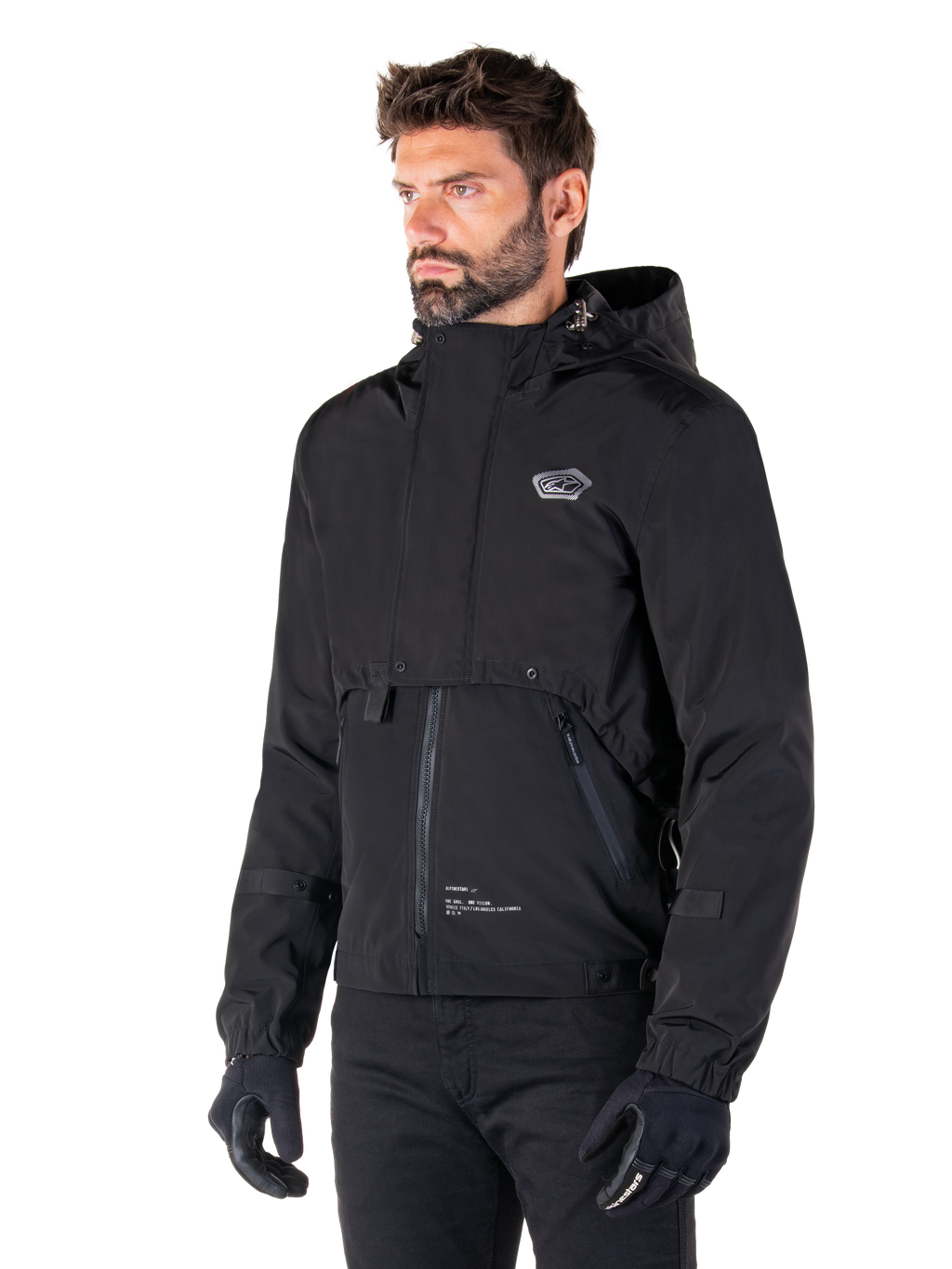 Alpinestars Mootant Waterproof Parka, zwarte modulaire motorjas, gedragen door een man met zwarte handschoenen en broek, voorzien van een capuchon, ritssluiting aan de voorzijde en gelaagd utilitair ontwerp, strakke urban esthetiek met logo op de borst.