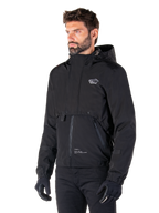 Alpinestars Mootant Waterproof Parka, zwarte modulaire motorjas, gedragen door een man met zwarte handschoenen en broek, voorzien van een capuchon, ritssluiting aan de voorzijde en gelaagd utilitair ontwerp, strakke urban esthetiek met logo op de borst.
