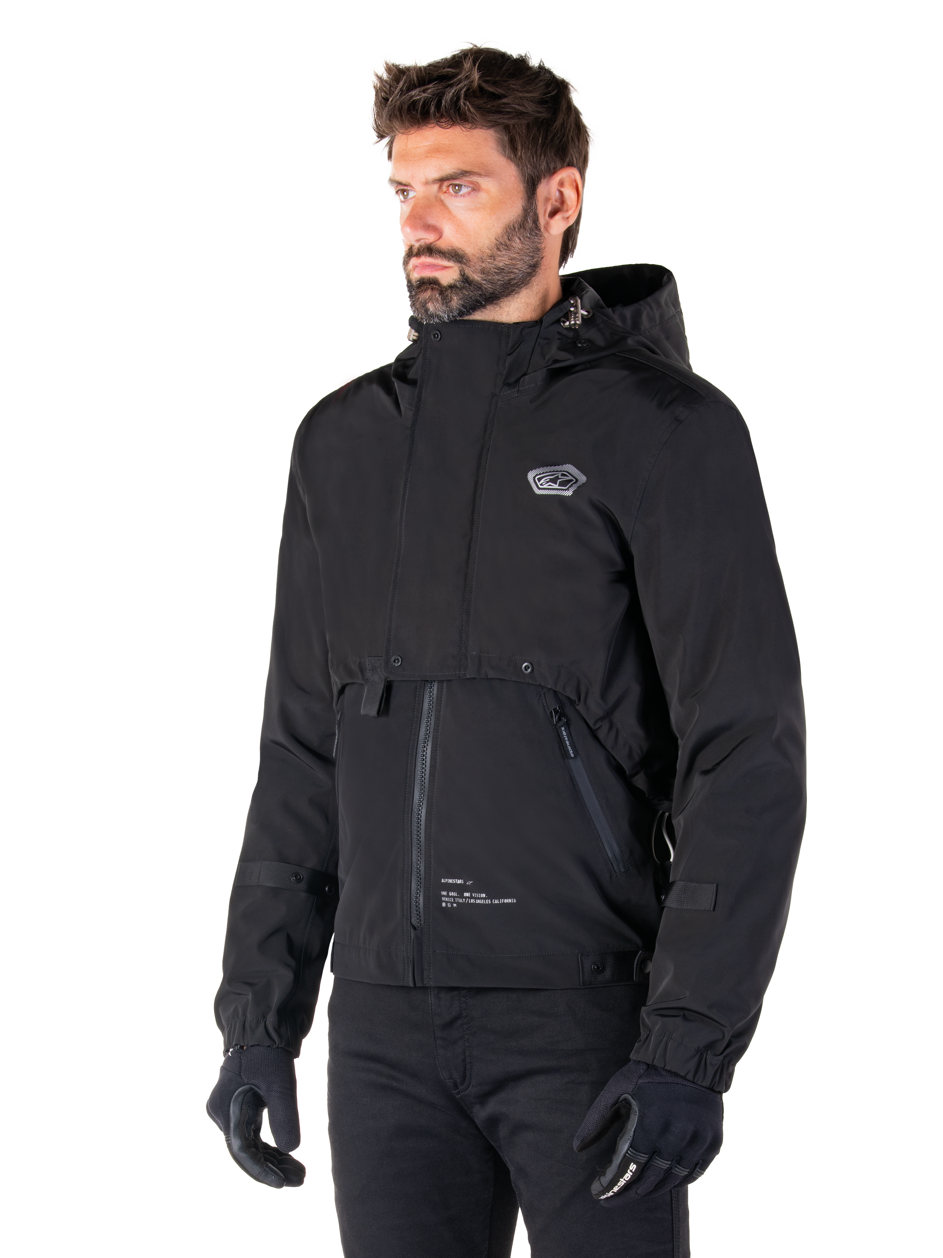 Alpinestars Mootant Waterproof Parka, zwarte modulaire motorjas, gedragen door een man met zwarte handschoenen en broek, voorzien van een capuchon, ritssluiting aan de voorzijde en gelaagd utilitair ontwerp, strakke urban esthetiek met logo op de borst.