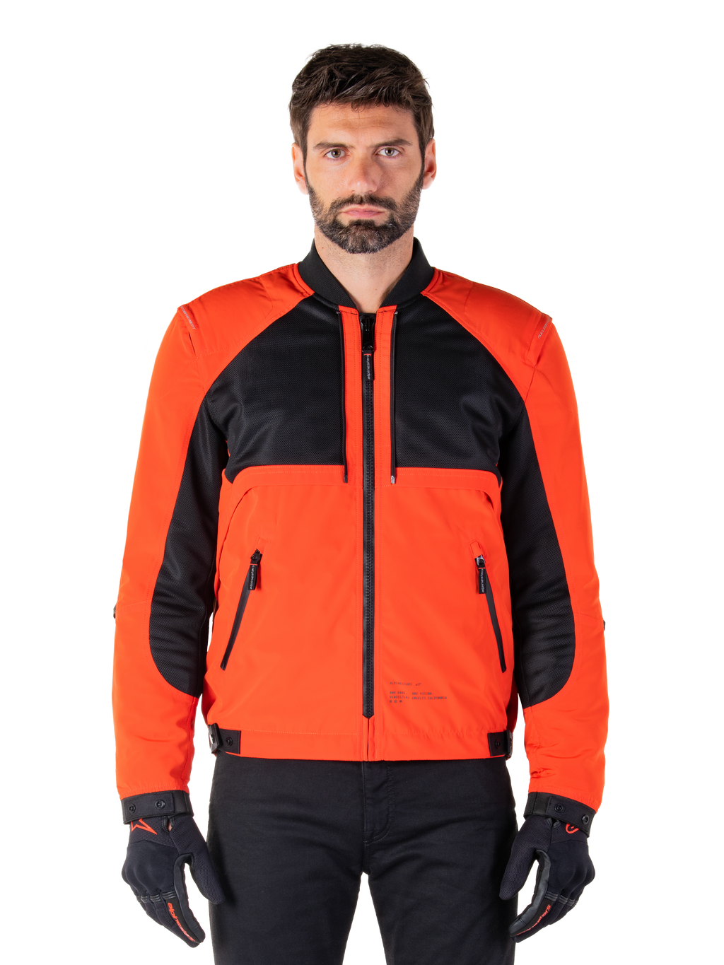 Alpinestars Mootant Waterproof Parka in Spicy Orange en zwart, modulaire motorjas getoond in een bomberjack-configuratie op een mannelijk model, met zwarte mesh panelen op de bovenborst, feloranje torso en mouwen, ritssluiting aan de voorzijde en een geribbelde kraag, gecombineerd met zwarte handschoenen en broek