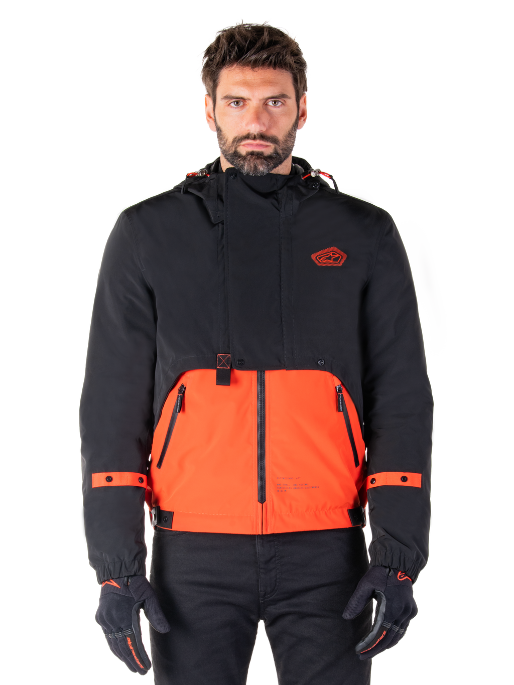 Alpinestars Mootant Waterproof Parka, modulaire motorjas in Spicy Orange en zwart, gedragen door een model met bijpassende handschoenen, voorzien van een ontwerp met capuchon en tweekleurige panelen, ritssluiting aan de voorzijde en verstelbare manchetten, urban contemporary motorkleding