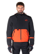 Alpinestars Mootant Waterproof Parka, modulaire motorjas in Spicy Orange en zwart, gedragen door een model met bijpassende handschoenen, voorzien van een ontwerp met capuchon en tweekleurige panelen, ritssluiting aan de voorzijde en verstelbare manchetten, urban contemporary motorkleding