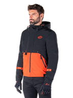 Man draagt de Alpinestars Mootant Waterproof Parka in Spicy Orange en Black, een modulaire motorjas met een zwarte bovenlaag met capuchon en een feloranje ondergedeelte, gecombineerd met zwarte motorhandschoenen en broek.