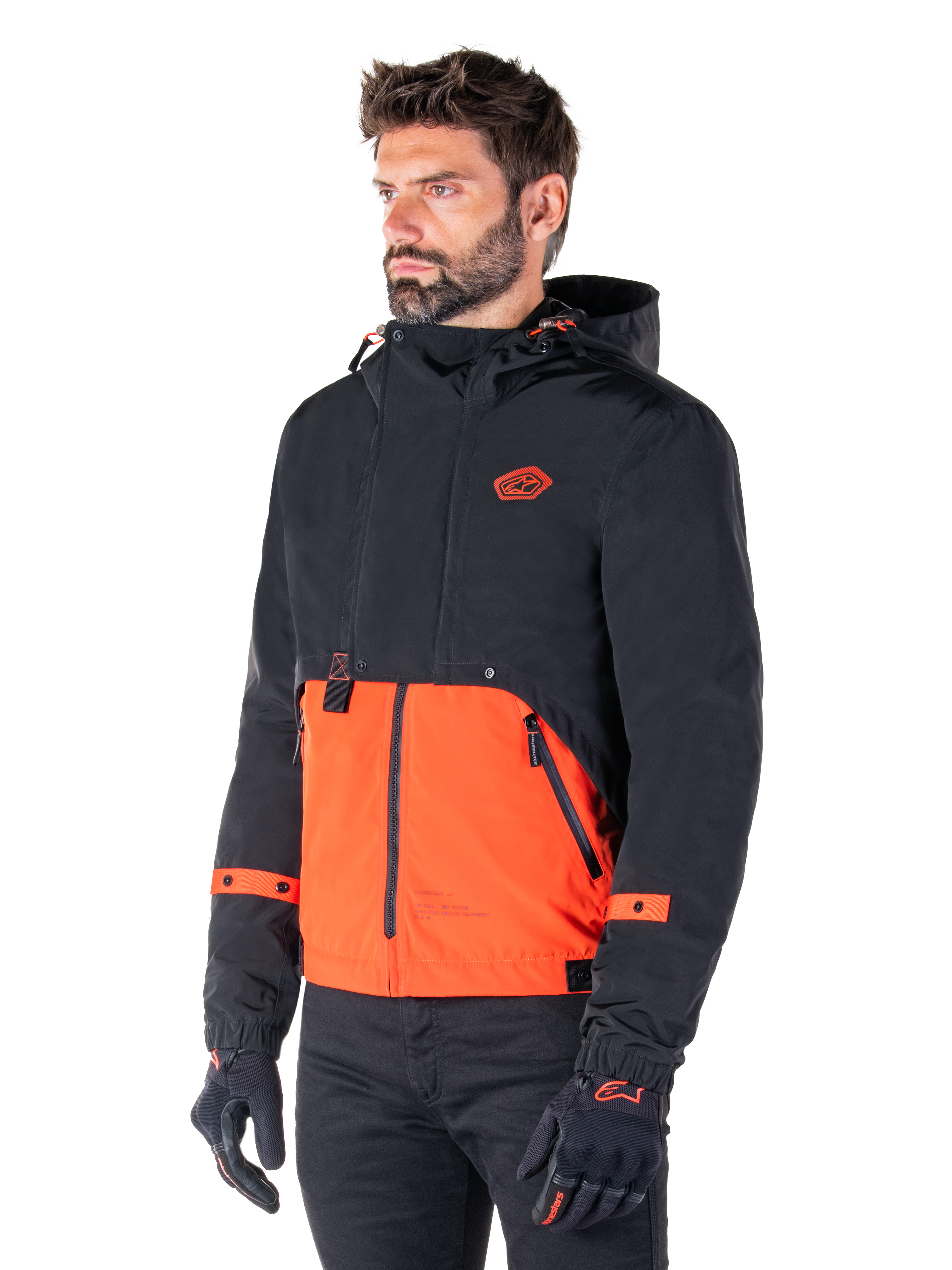 Man draagt de Alpinestars Mootant Waterproof Parka in Spicy Orange en Black, een modulaire motorjas met een zwarte bovenlaag met capuchon en een feloranje ondergedeelte, gecombineerd met zwarte motorhandschoenen en broek.