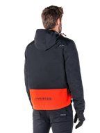 Alpinestars Mootant Waterproof Parka, Spicy Orange en zwart, achteraanzicht op een mannelijk model, voorzien van een zwart bovengedeelte met capuchon en een feloranje ondergedeelte met MO.ST.EQ.-branding, urban contemporary motorjas gedragen met een zwarte broek en handschoenen