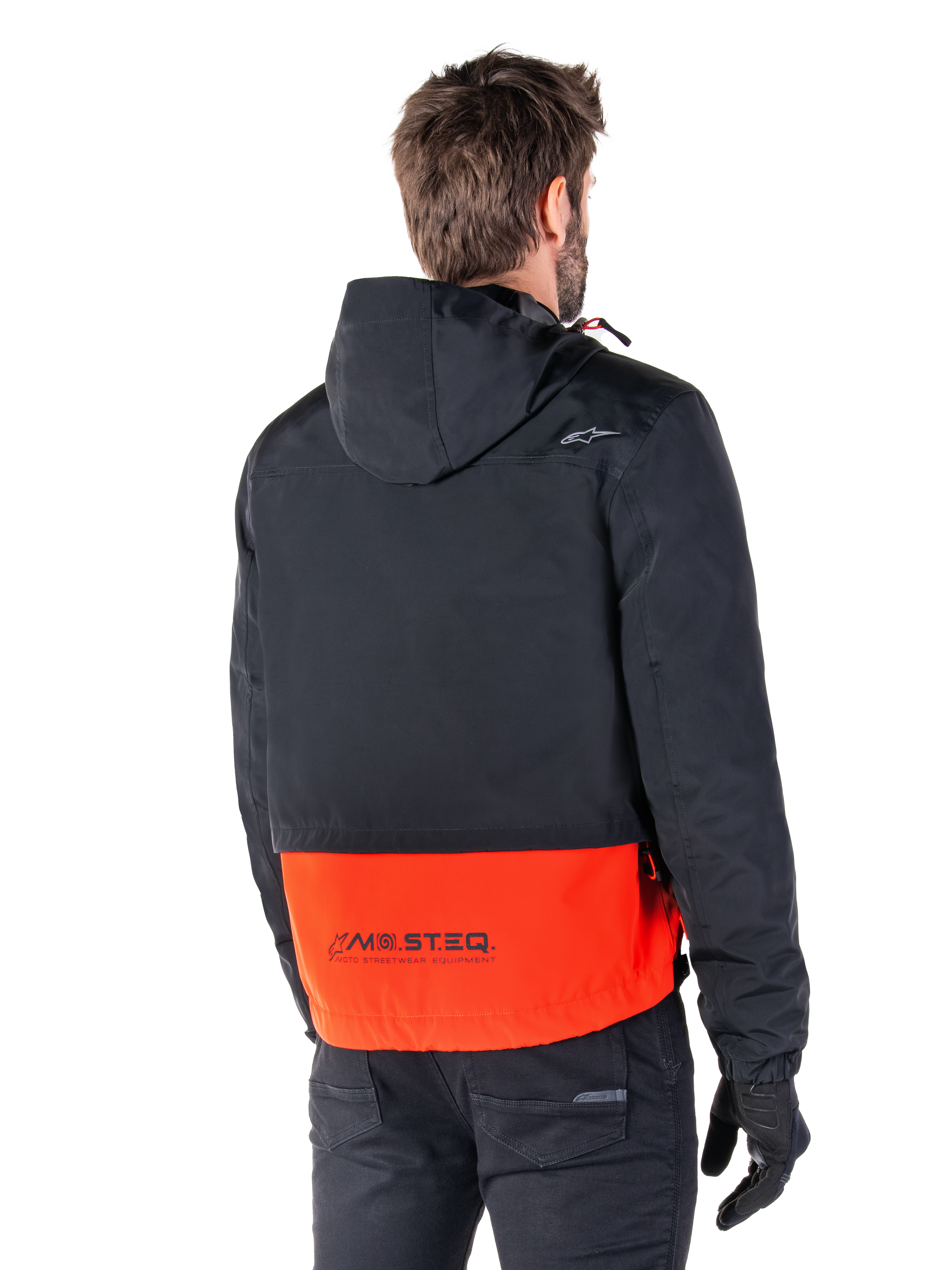 Alpinestars Mootant Waterproof Parka, Spicy Orange en zwart, achteraanzicht op een mannelijk model, voorzien van een zwart bovengedeelte met capuchon en een feloranje ondergedeelte met MO.ST.EQ.-branding, urban contemporary motorjas gedragen met een zwarte broek en handschoenen
