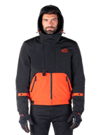 Alpinestars Mootant Waterproof Parka, motorjas, Spicy Orange en zwart, modulair all-weather ontwerp met capuchon, ritszakken aan de voorkant en oranje banddetails op de mouwen, gedragen door een model met zwarte handschoenen en broek