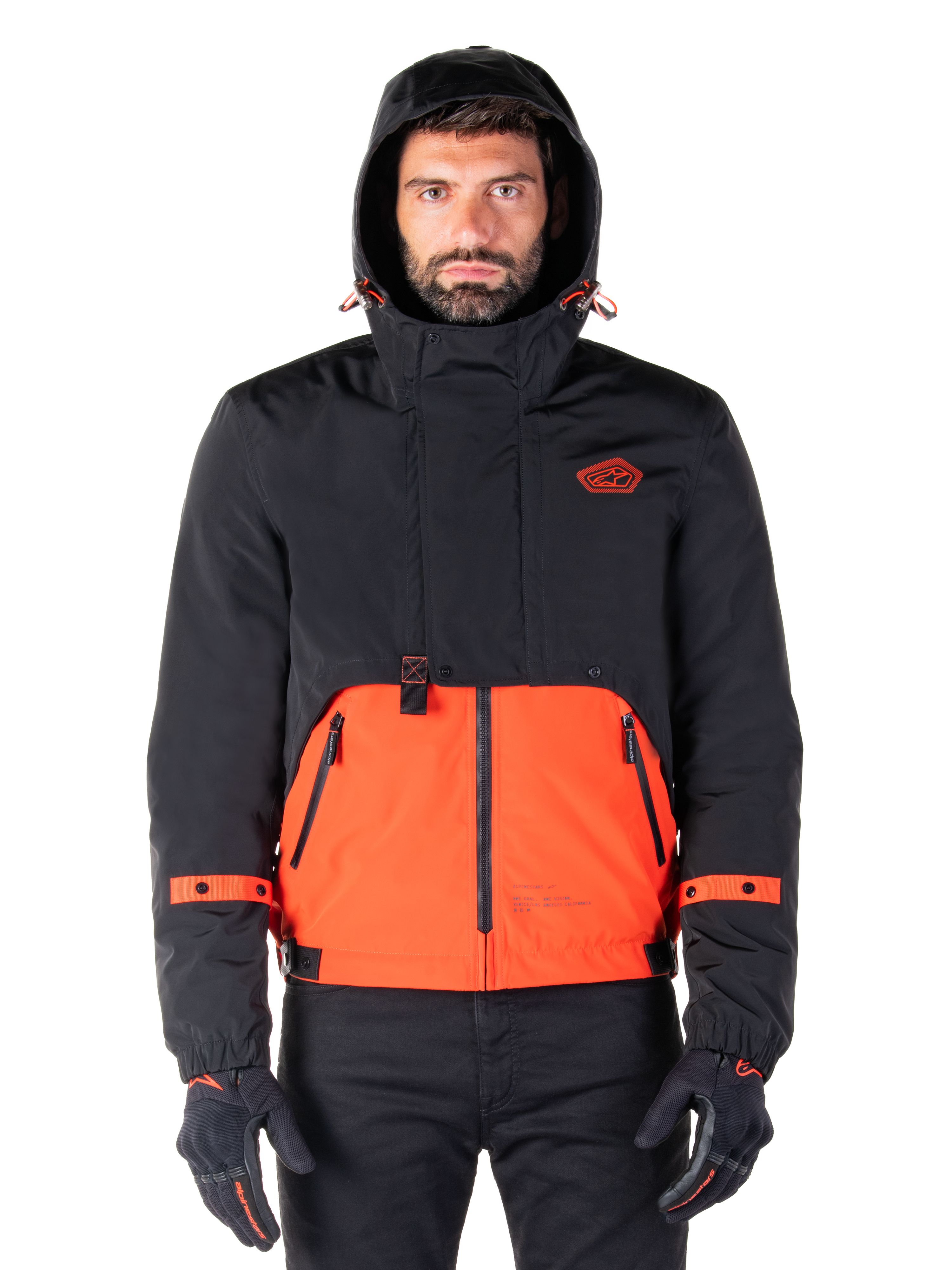 Alpinestars Mootant Waterproof Parka, motorjas, Spicy Orange en zwart, modulair all-weather ontwerp met capuchon, ritszakken aan de voorkant en oranje banddetails op de mouwen, gedragen door een model met zwarte handschoenen en broek
