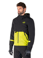 Alpinestars Mootant Waterproof Parka in Lime Punch Black, modulaire motorjas gedragen door een man met zwarte motorhandschoenen en broek, met een zwart bovenstuk en mouwen en een opvallend limoengroen paneel aan de onderkant, ontwerp met capuchon en een utilitaire, militair geïnspireerde esthetiek