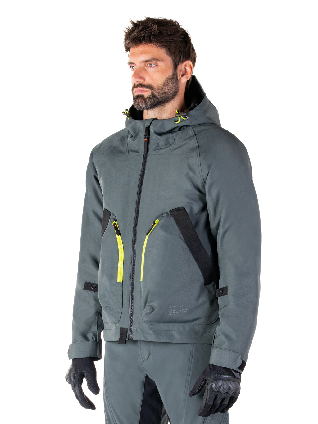 Alpinestars Morush Waterproof Parka, Urban Gray motorjas, ontwerp met capuchon en felgele ritsaccenten, zwarte zakdetails, gedragen met bijpassende grijze motorbroek en zwarte handschoenen, eigentijdse op streetwear geïnspireerde motorkleding.