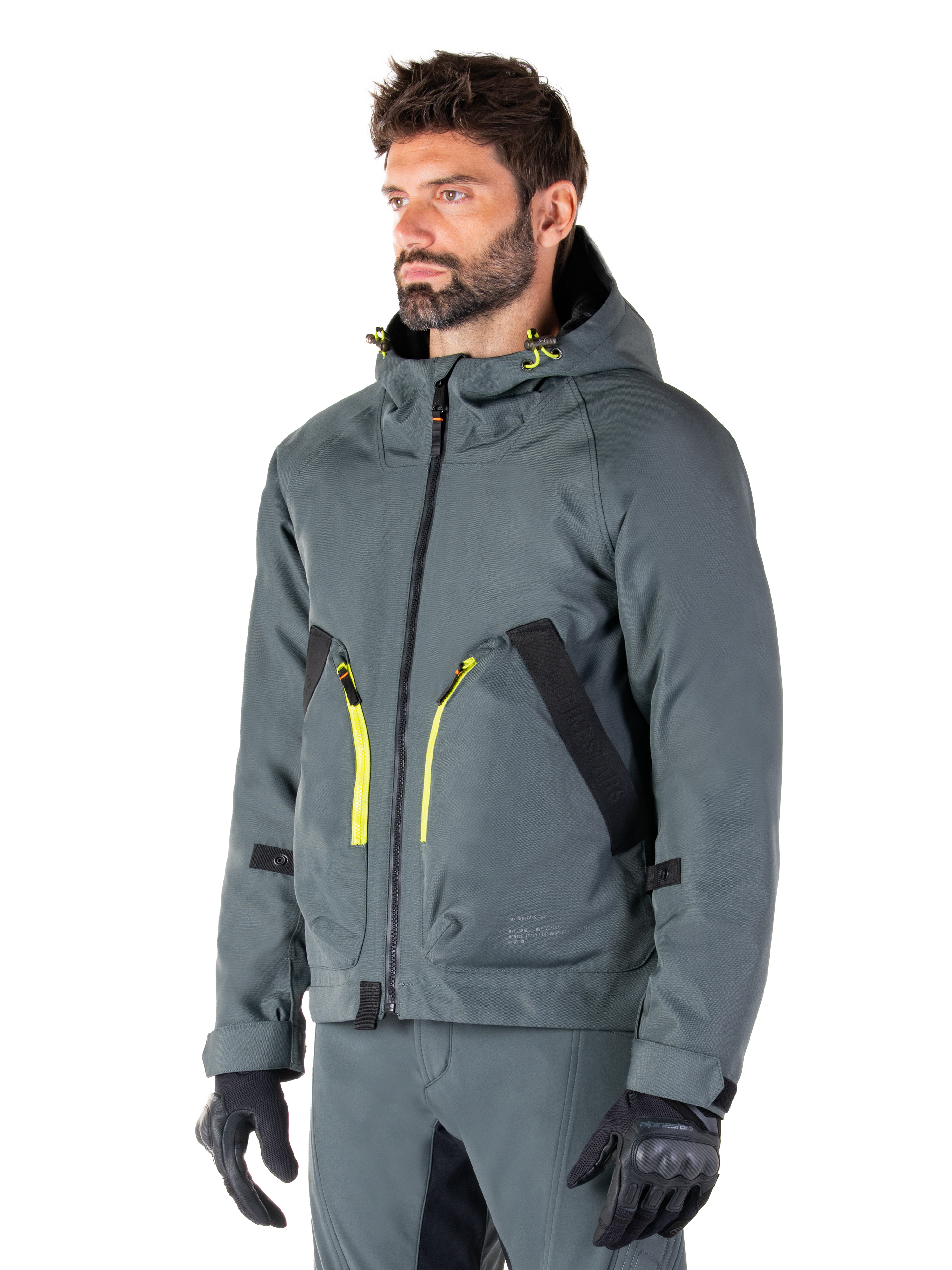 Alpinestars Morush Waterproof Parka, Urban Gray motorjas, ontwerp met capuchon en felgele ritsaccenten, zwarte zakdetails, gedragen met bijpassende grijze motorbroek en zwarte handschoenen, eigentijdse op streetwear geïnspireerde motorkleding.
