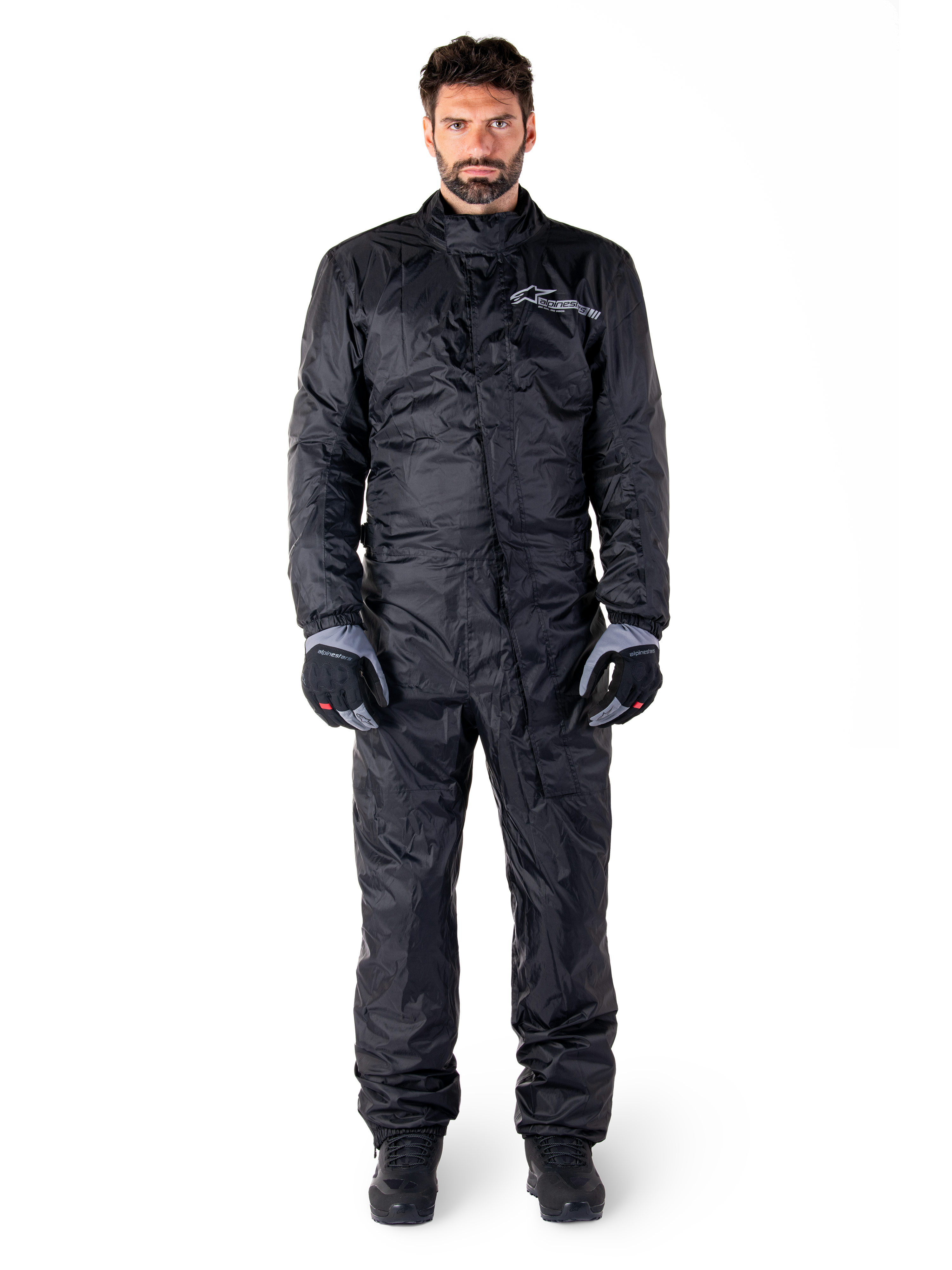 Alpinestars Hurricane Rain V2 Suit, volledig motorregenpak in het zwart, waterdichte beschermende kleding met hoge kraag en stormsluiting aan de voorzijde, voorzien van een wit logo op de borst, gedragen met zwart-grijze handschoenen en laarzen