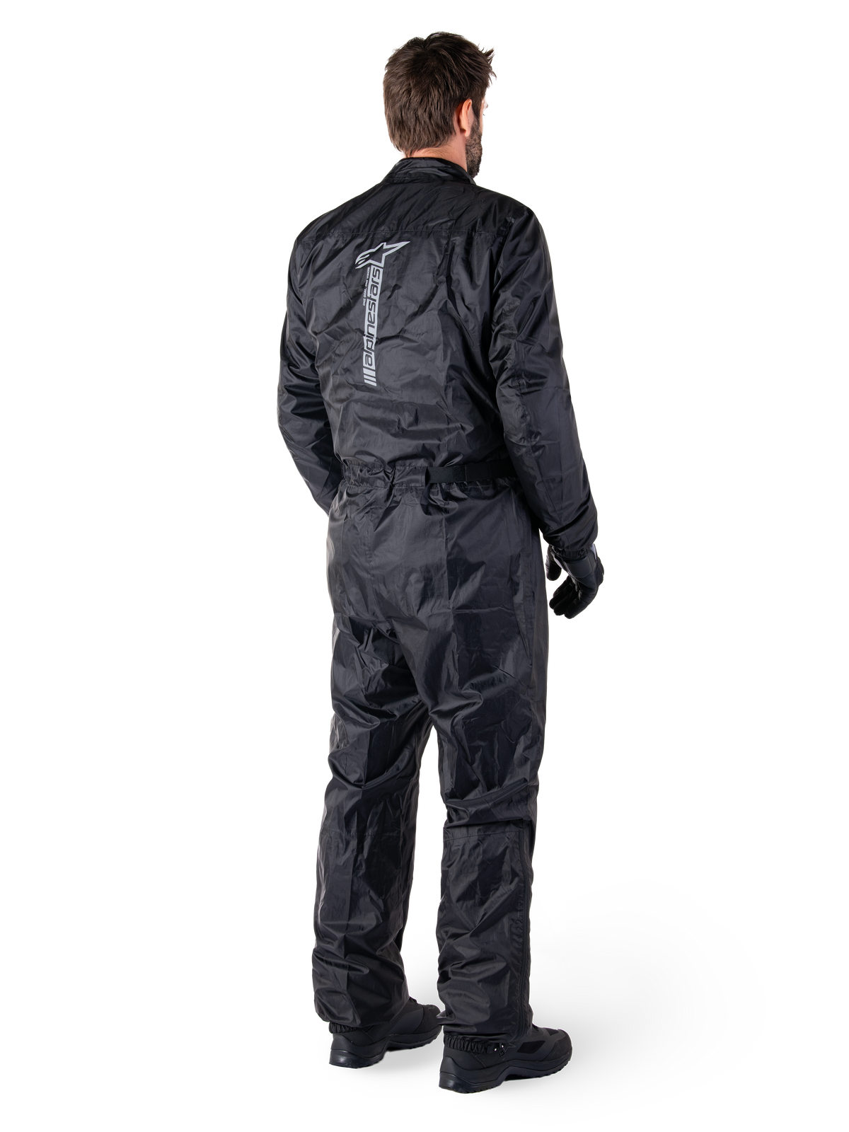 Alpinestars Hurricane Rain V2-pak, zwart eendelig waterdicht motorregenpak, achteraanzicht met groot verticaal reflecterend logo, lichtgewicht windbestendige stof, gedragen met zwarte handschoenen en laarzen