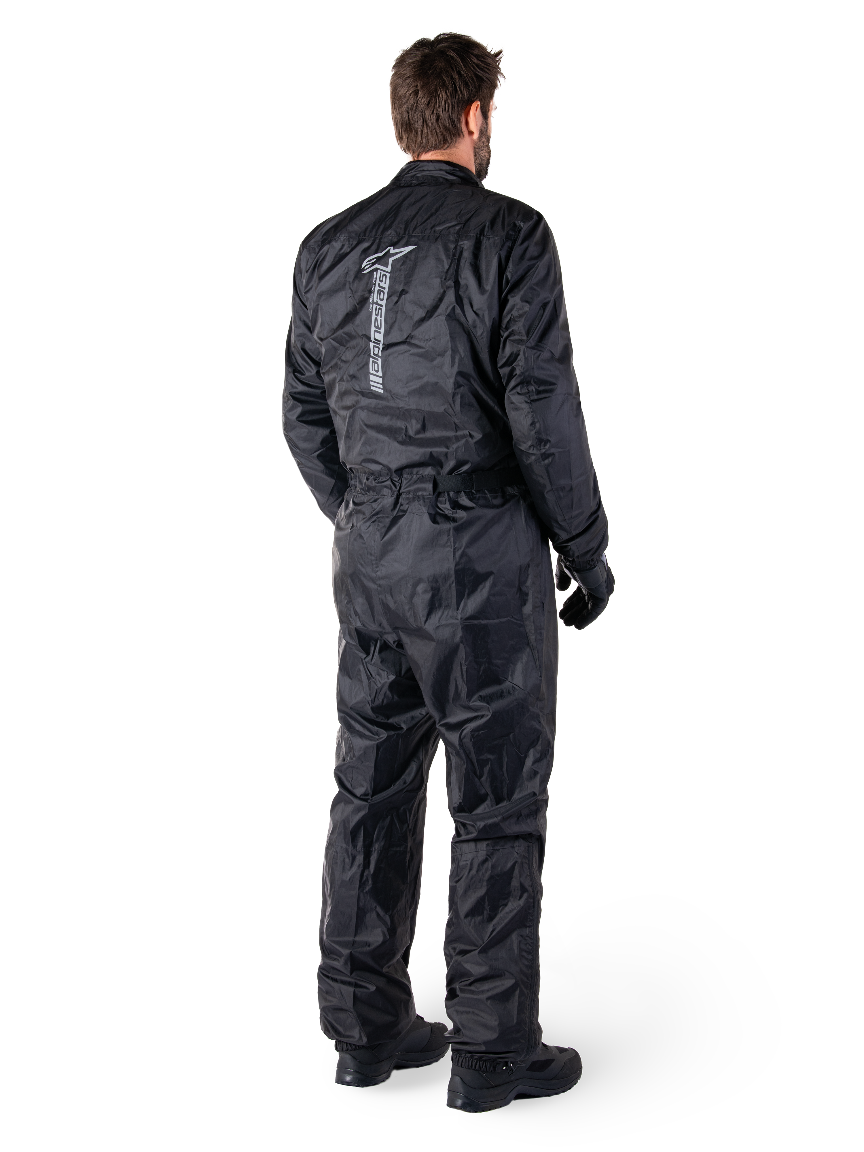 Alpinestars Hurricane Rain V2-pak, zwart eendelig waterdicht motorregenpak, achteraanzicht met groot verticaal reflecterend logo, lichtgewicht windbestendige stof, gedragen met zwarte handschoenen en laarzen
