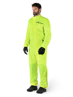 Alpinestars Hurricane Rain V2 Pak, eendelig waterdicht motorregenpak, fluoriserend geel, gedragen met zwarte handschoenen en laarzen, voorzien van een zwart Alpinestars-logo op de borst en reflecterende details op de benen