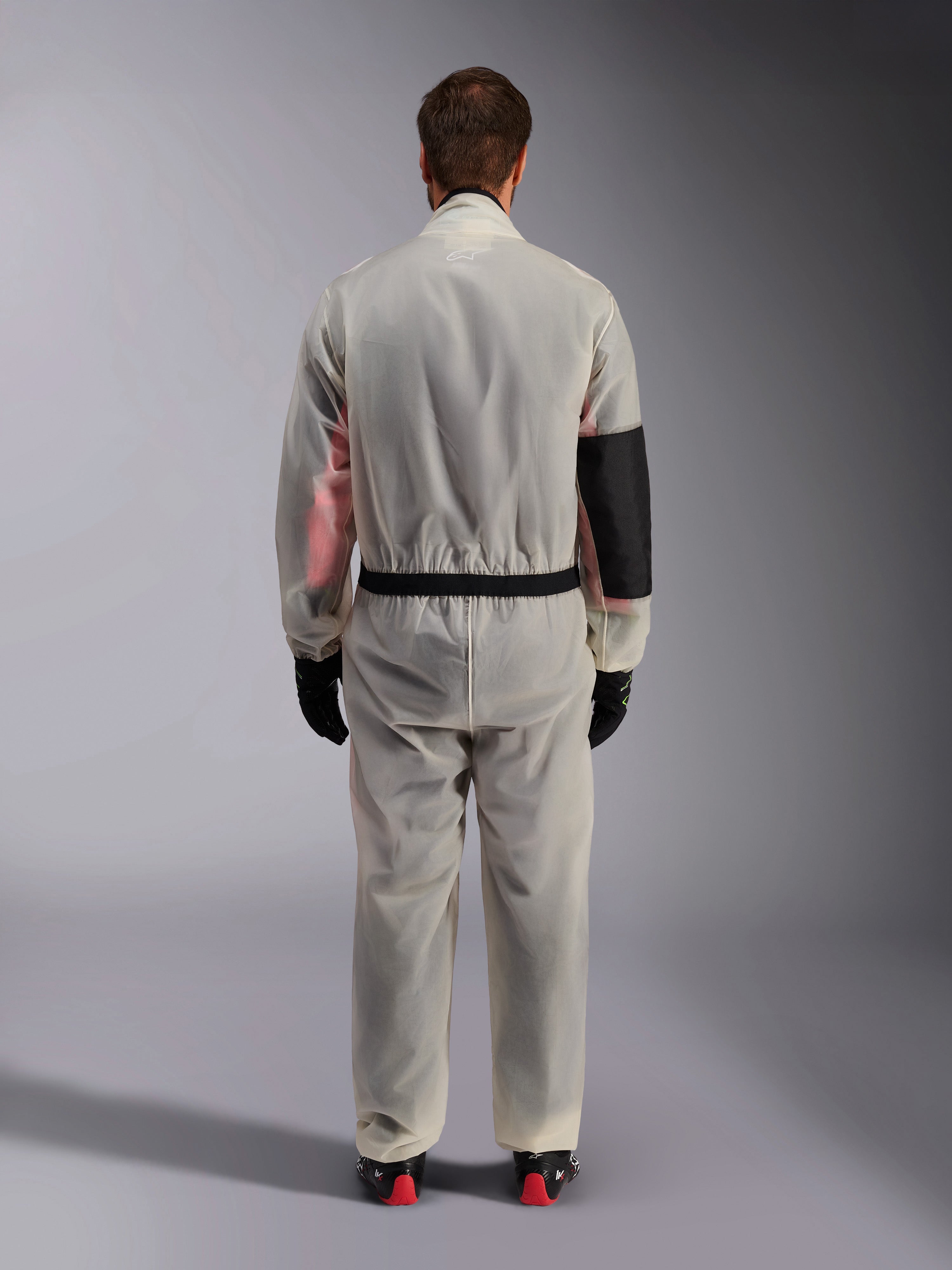 Alpinestars Kart Rain Suit, Karting Oversuit, Translucent Clear, back view showing a reinforced black panel on the right elbow for heat protection, elasticized waist with black trim, see-through material revealing a racing suit underneath, paired with black gloves and racing boots.