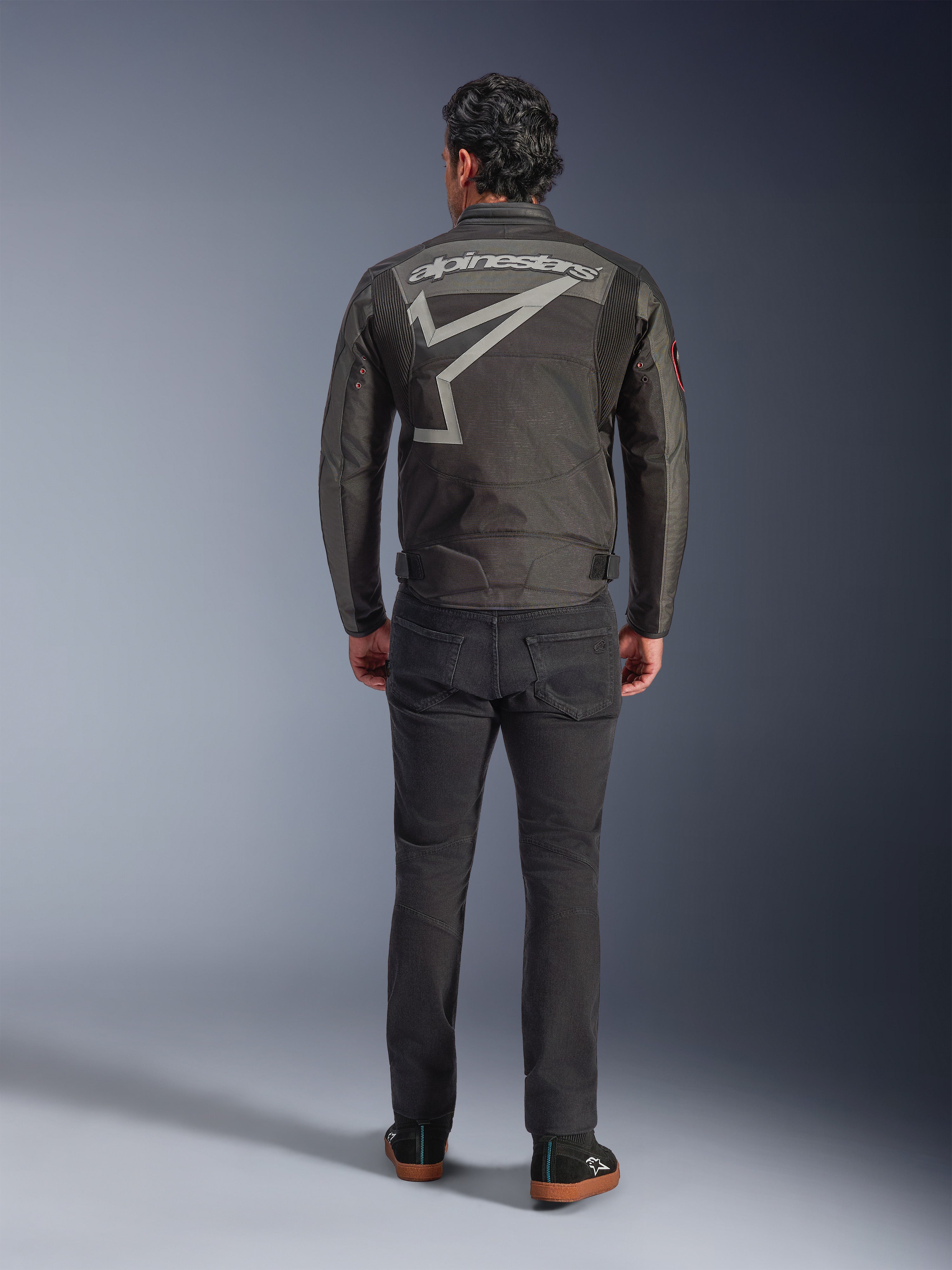 Alpinestars Axton Jacket, motorjas, Black Anthracite Red Tangerine, achteraanzicht met groot grijs Alpinestars-logo en stergrafiek, accordeonpanelen op de schouders, gedragen met zwarte broek en casual zwarte schoenen