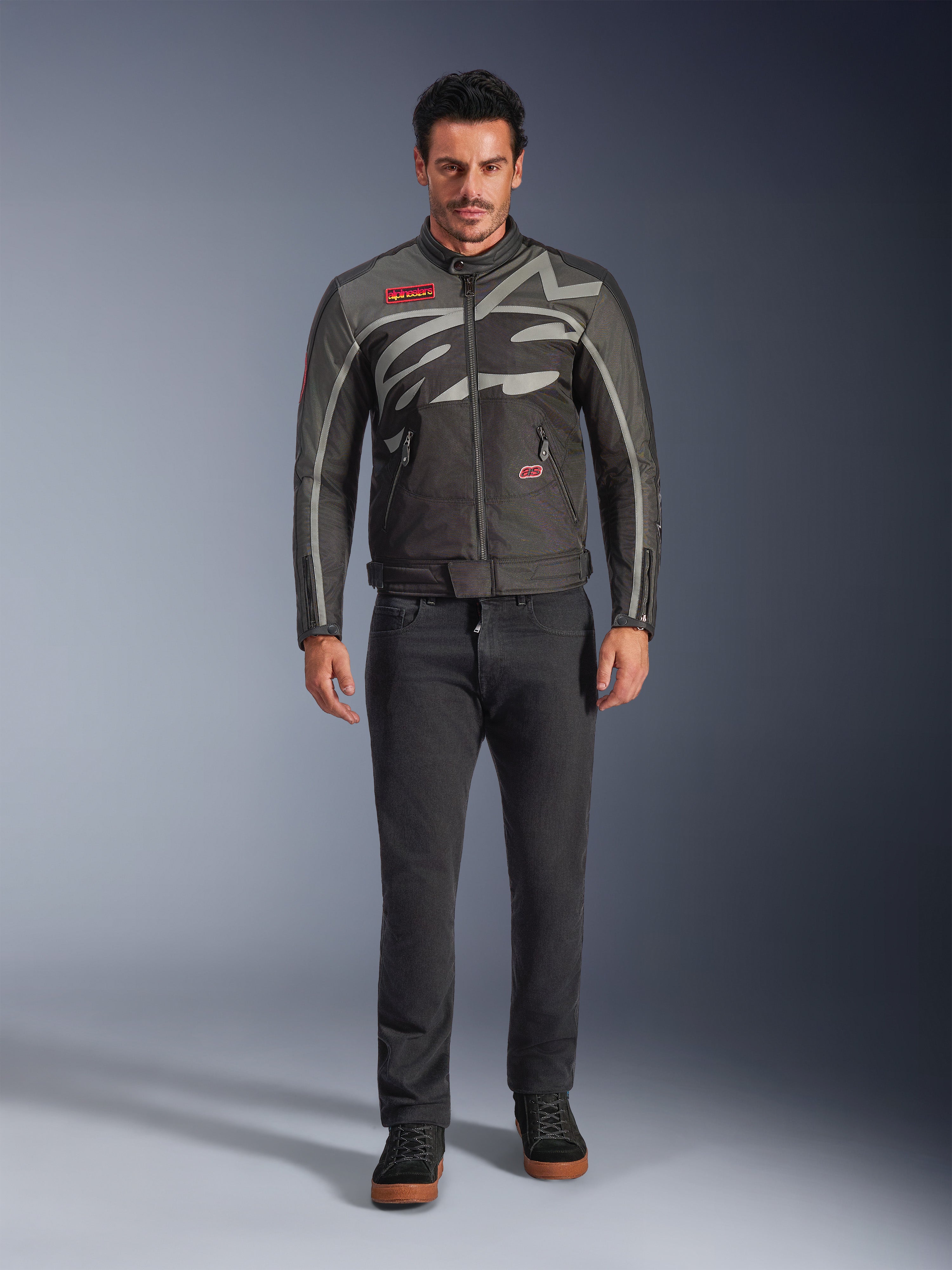 Alpinestars Axton jas, motorjas, Black Anthracite Red Tangerine, gedragen door een model met een donkere broek, voorzien van een rondom lopend grijs Alpinestars-logo, rode merkpatches en een waterbestendige textiele buitenlaag voor stadsritten