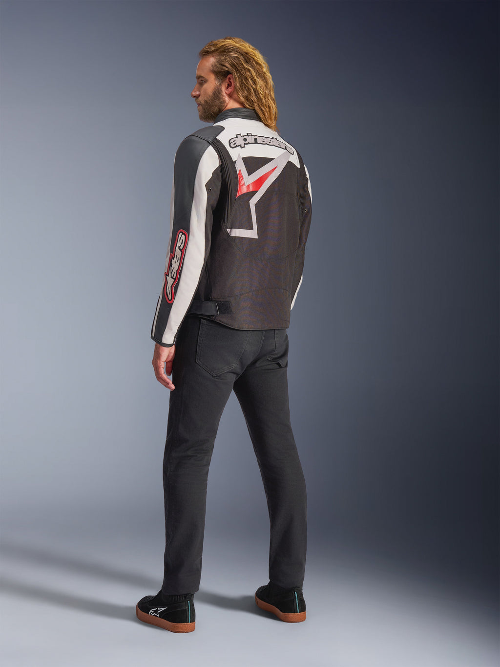 Alpinestars Axton Jas, motorjas, zwart, gebroken wit en felrood, achteraanzicht gedragen door mannelijk model, voorzien van groot Alpinestars-sterlogo en woordmerk op de rug, vintage racing-geïnspireerd ontwerp met accordeonpanelen, gecombineerd met zwarte broek en sneakers