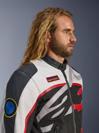Man met lang haar en baard draagt het Alpinestars Axton Jacket, motorjas, zwart, off-white en felrood, vintage racing-geïnspireerd ontwerp met merkpatches en rondom lopend logo, close-up profielaanzicht, urban contemporary stijl