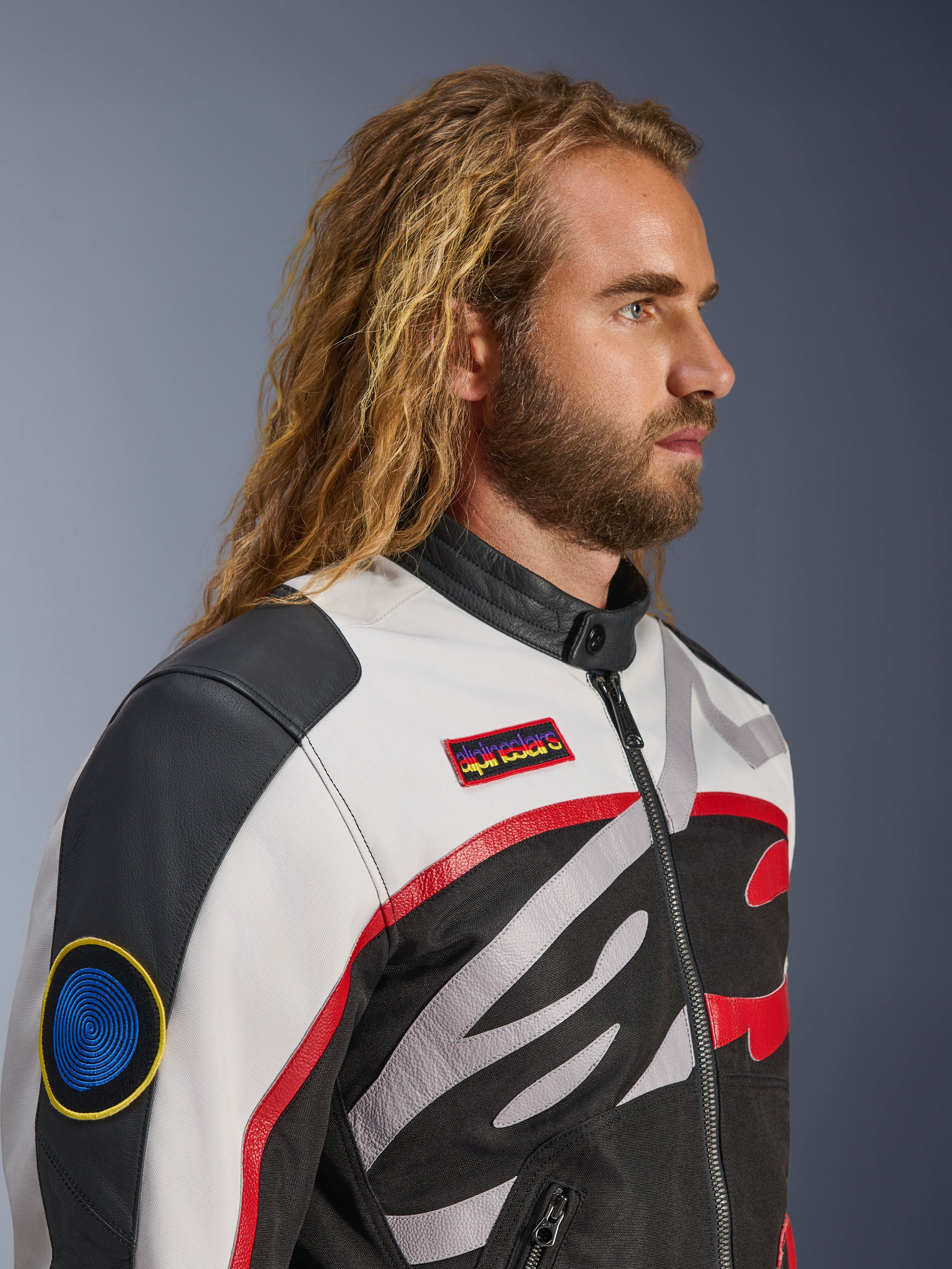 Man met lang haar en baard draagt het Alpinestars Axton Jacket, motorjas, zwart, off-white en felrood, vintage racing-geïnspireerd ontwerp met merkpatches en rondom lopend logo, close-up profielaanzicht, urban contemporary stijl