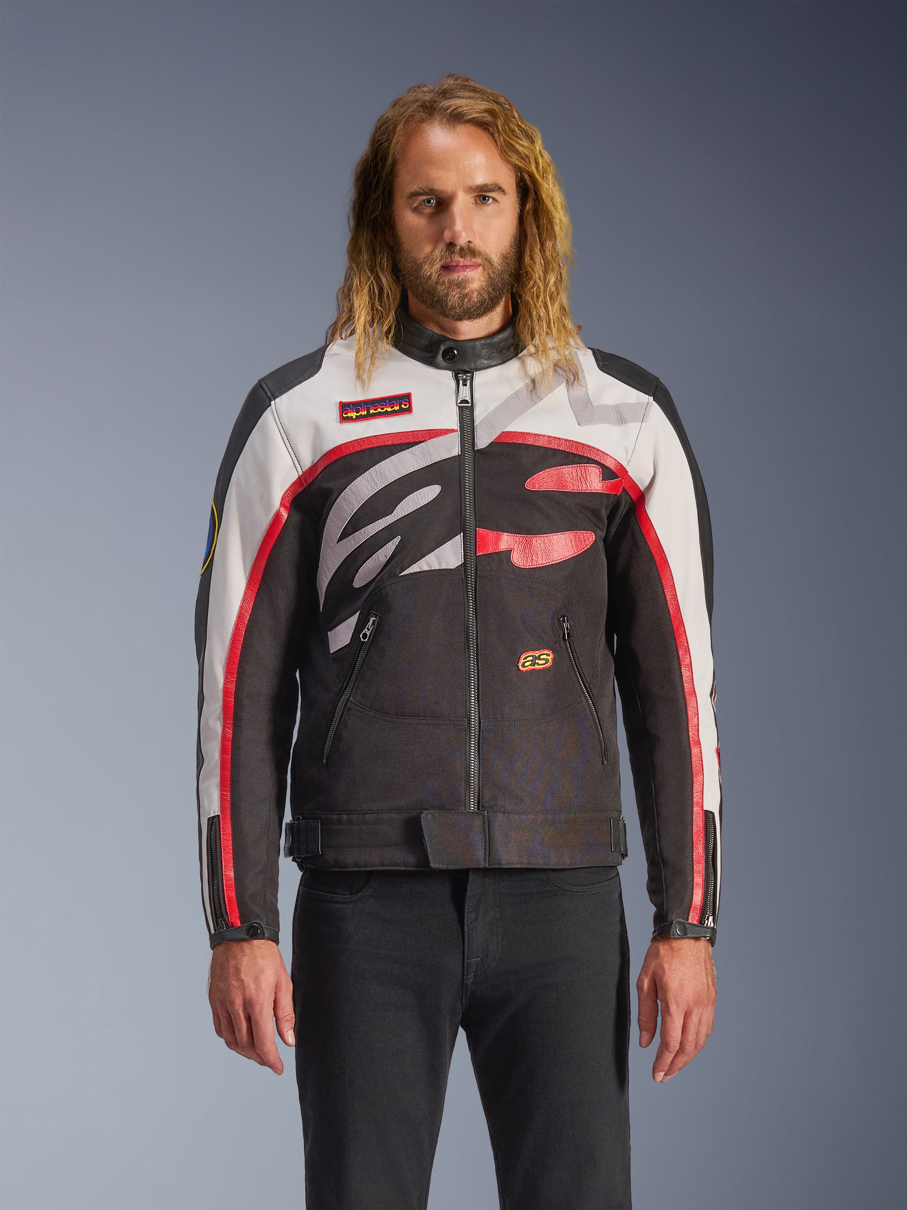 Alpinestars Axton Jas, motorjas, zwart, off-white en felrood, voorzien van opvallende abstracte patronen op de borst en een rondom lopend logo, mouwen met rode strepen, vintage-geïnspireerd ontwerp met een minimalistische kraag, gedragen door een model met zwarte jeans.