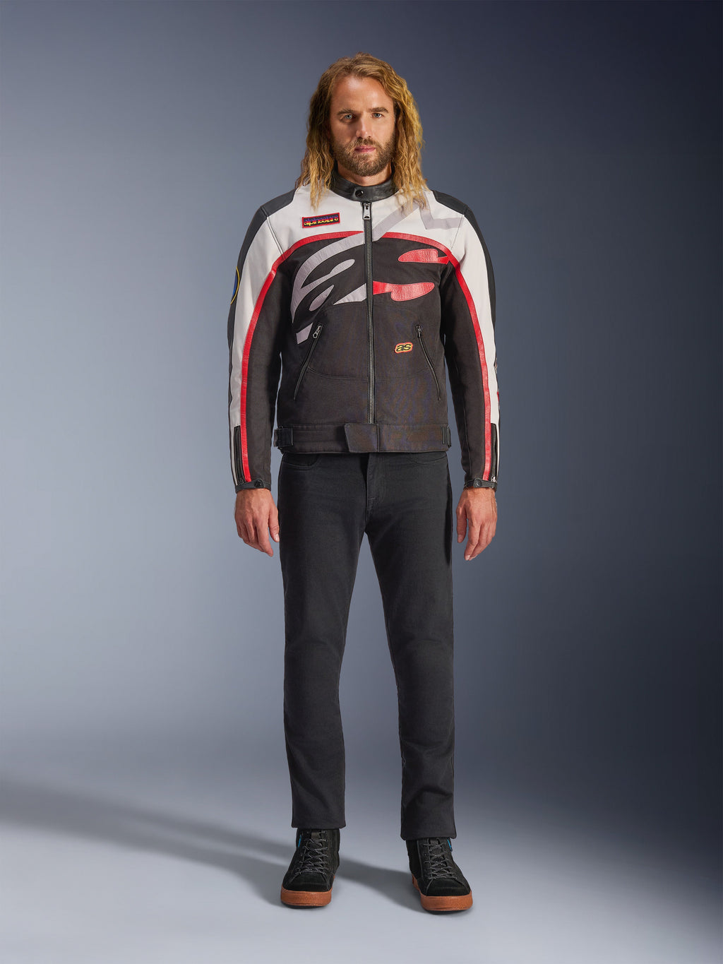 Alpinestars Axton Jacket, motorjas, zwart, gebroken wit en felrood, gedragen door een model met een zwarte broek en sneakers, voorzien van een opvallend rondom lopend logo en een vintage op de racerij geïnspireerd ontwerp