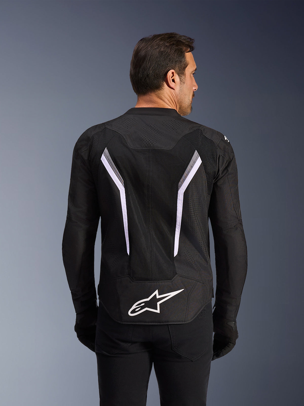 Alpinestars AST V3 Air Jacket, motorjas, zwart met witte en donkergrijze accenten, achteraanzicht met uitgebreide mesh ventilatiepanelen, grote witte V-vormige strepen en wit Alpinestars-logo op de onderrug, gedragen met zwarte handschoenen en broek