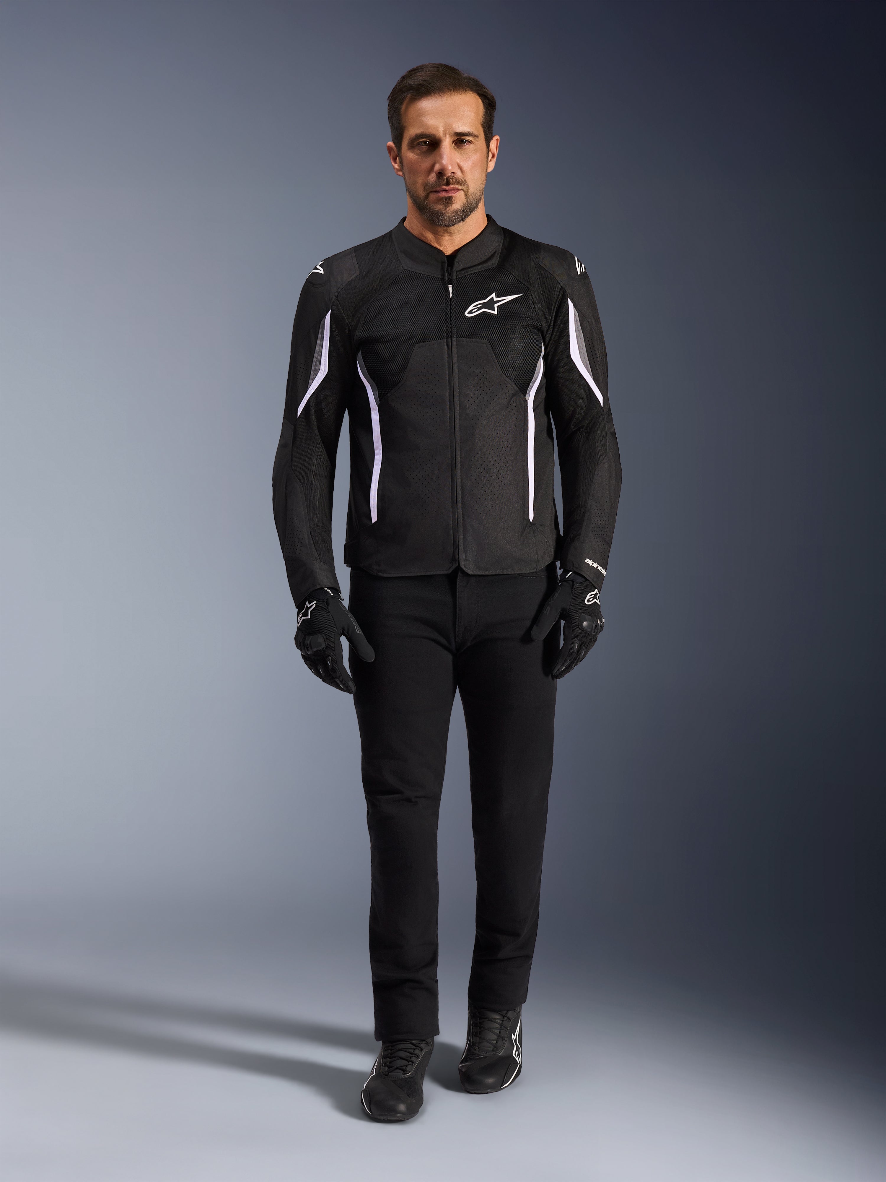 Mannelijk model draagt het Alpinestars AST V3 Air Jacket in zwart met witte accenten, voorzien van mesh ventilatiepanelen en een sportieve pasvorm, gecombineerd met een zwarte technische motorbroek, handschoenen en laarzen, vooraanzicht, technische motoruitrusting voor rijden bij warm weer.