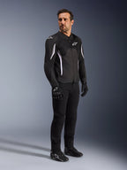 Alpinestars AST V3 Air Jacket, zwart met witte accenten en logo's, lichtgewicht motorjas met mesh-ventilatie, gedragen door een man met zwarte motorbroek, handschoenen en schoenen, technische zomeruitrusting voor sport- en straatgebruik.