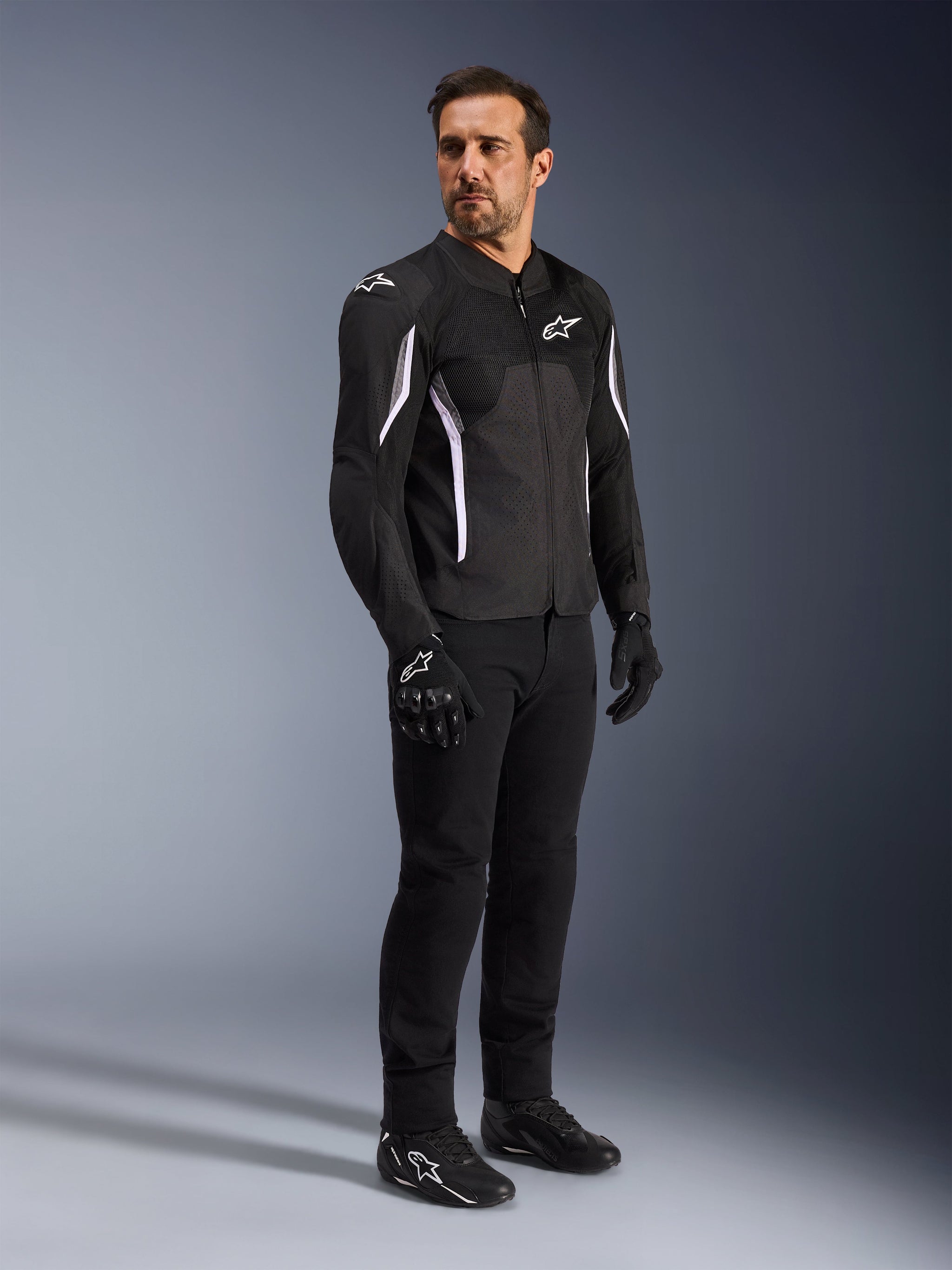Alpinestars AST V3 Air Jacket, zwart met witte accenten en logo's, lichtgewicht motorjas met mesh-ventilatie, gedragen door een man met zwarte motorbroek, handschoenen en schoenen, technische zomeruitrusting voor sport- en straatgebruik.