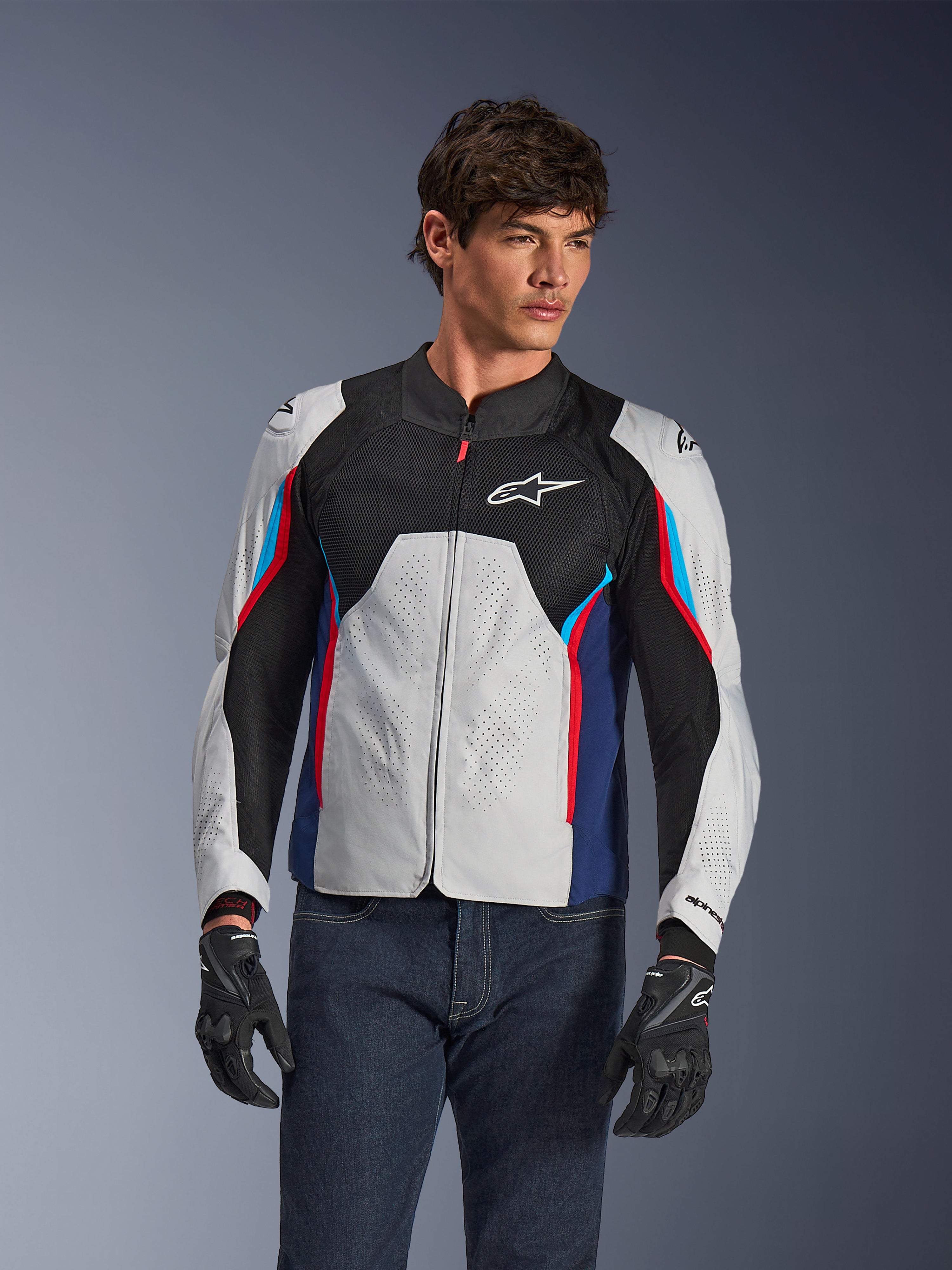Alpinestars AST V3 Air Jacket in Silver Deep Blue Bright Red, gedragen door een model met een donkere jeans en zwarte handschoenen, voorzien van geperforeerde zilveren panelen, zwarte mesh ventilatie en sportieve rode en blauwe accenten.