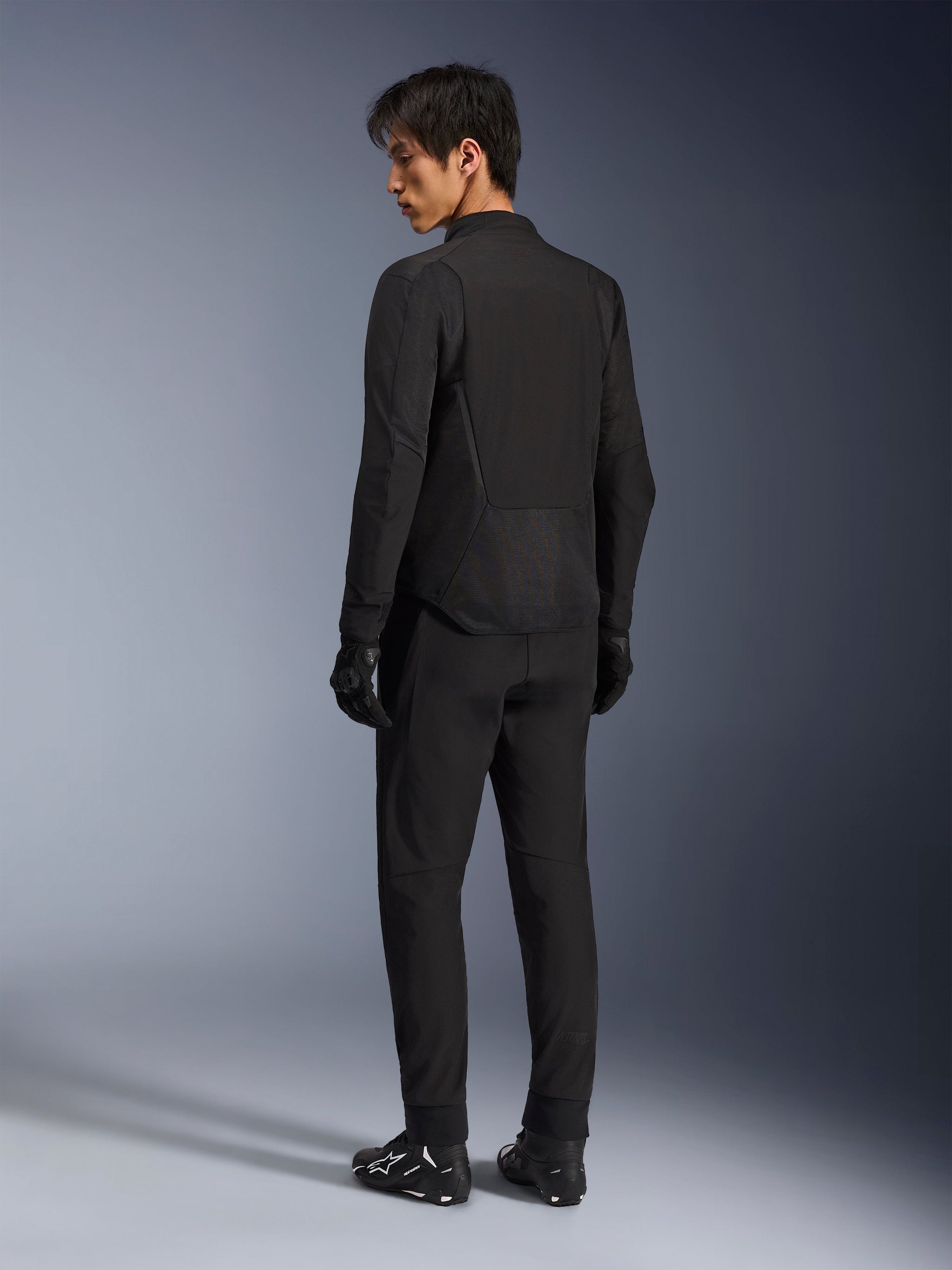 Alpinestars Aeroshell Airflow Jacket, zwart, lichtgewicht textiele motorjas, gedragen door model met bijpassende zwarte uitrusting, achteraanzicht met de nadruk op lasergeperforeerde ventilatiepanelen en sportieve pasvorm met verlengde achterkant.