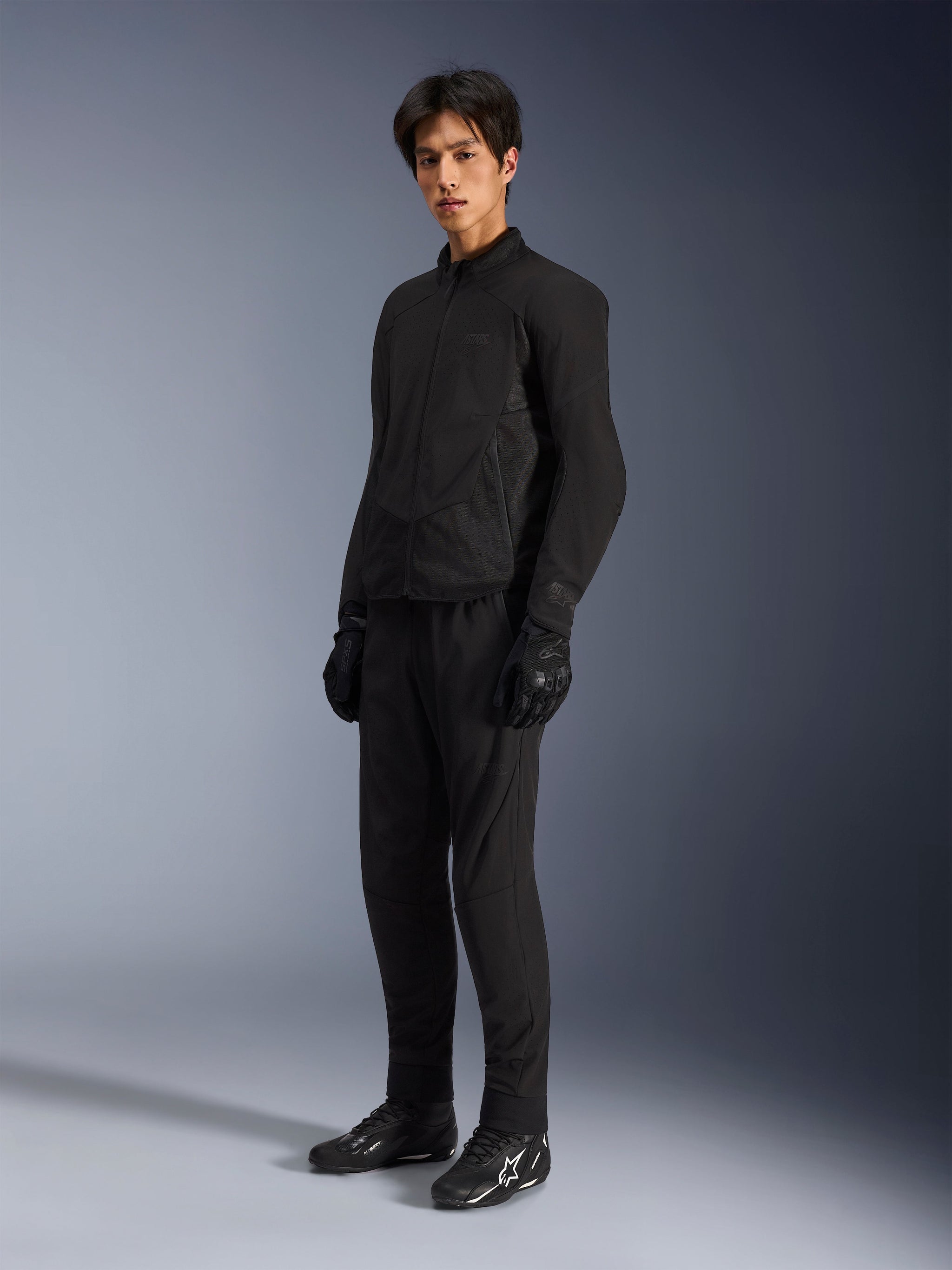 Alpinestars Aeroshell Airflow Jacket, Black Anthracite motorjas, voorzien van lasergeperforeerde ventilatie en meshpanelen voor optimale luchtstroom, gedragen door een mannelijk model met bijpassende zwarte motorbroek, handschoenen en zwarte laarzen met witte logo's, sportieve pasvorm voor wegprestaties