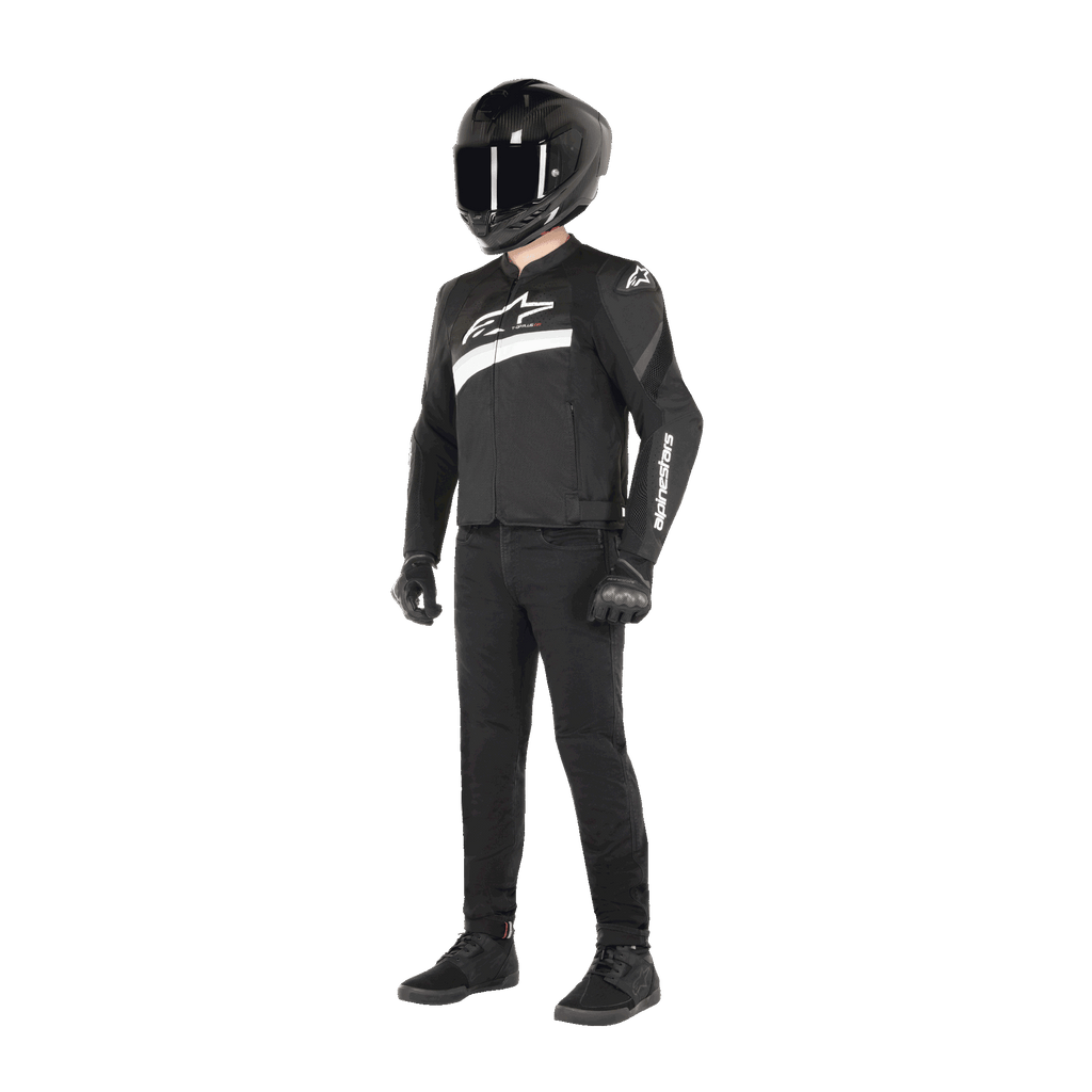 Motorrijder die de Alpinestars T-GP Plus R V4 Airflow Jas draagt in zwart en wit, textiele motorjas met mesh-panelen en witte Alpinestars-branding, gecombineerd met een zwarte helm, zwarte motorhandschoenen, een zwarte broek en zwarte schoenen