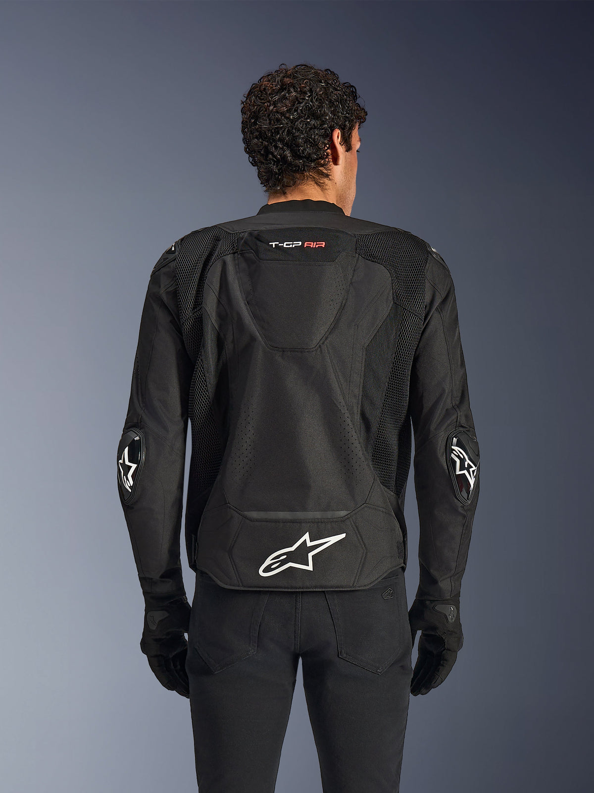 Alpinestars T-GP Air jas, zwart, achteraanzicht van een textiele motorjas, voorzien van een aerodynamische bult, grote mesh ventilatiepanelen en een wit Alpinestars-logo op de onderrug, gecombineerd met zwarte motorhandschoenen en broek