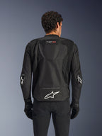 Alpinestars T-GP Air jas, zwart, achteraanzicht van een textiele motorjas, voorzien van een aerodynamische bult, grote mesh ventilatiepanelen en een wit Alpinestars-logo op de onderrug, gecombineerd met zwarte motorhandschoenen en broek