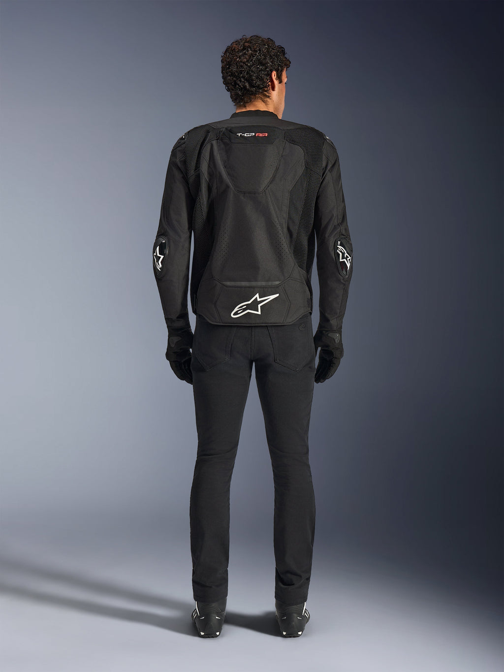Alpinestars T-GP Air Jacket, zwart, achteraanzicht van een man die de textiele motorjas draagt, voorzien van mesh ventilatiepanelen, een aerodynamische bult en witte Alpinestars-logo's op de onderrug en ellebogen, gedragen met een zwarte motorbroek en handschoenen