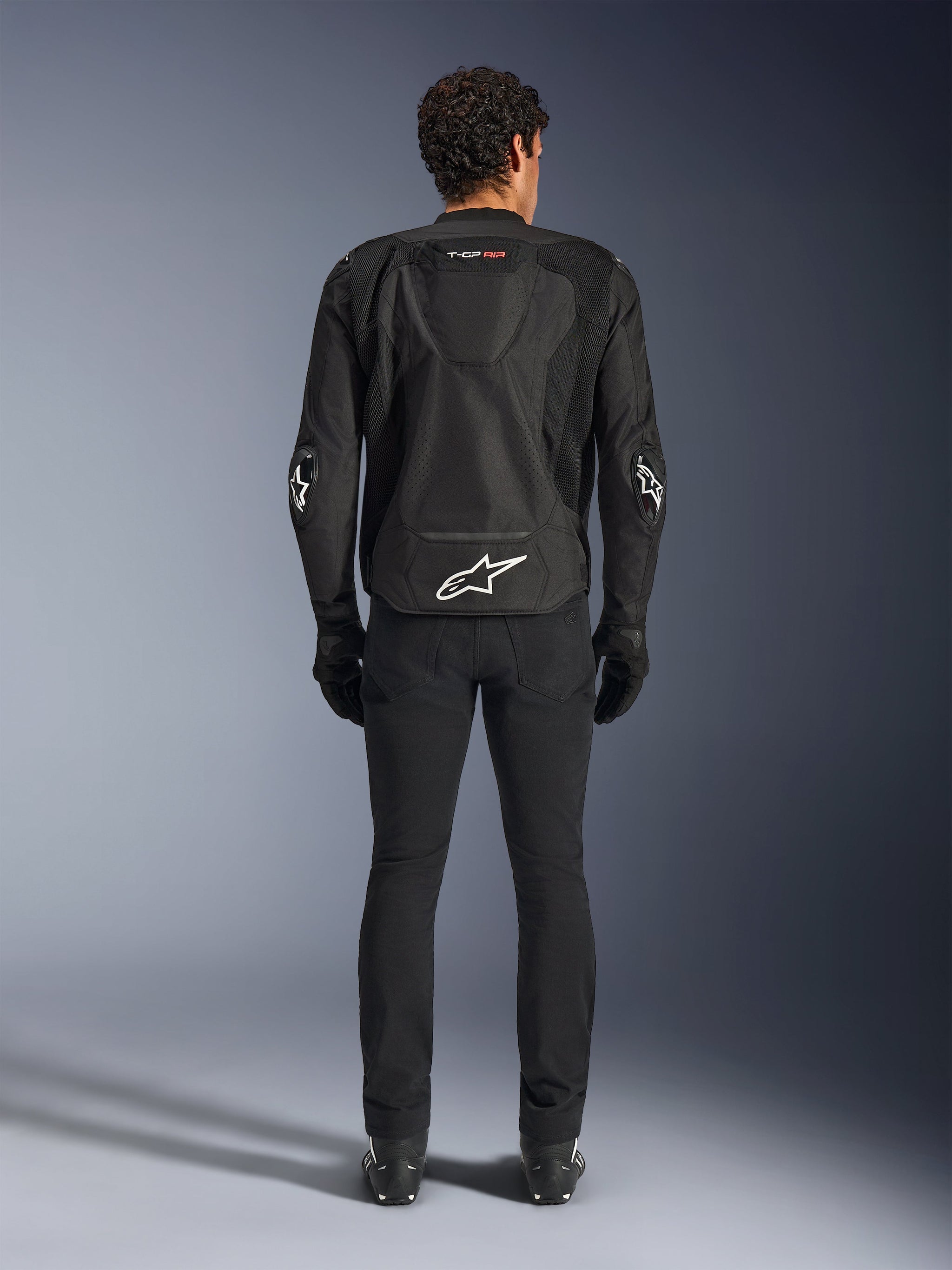 Alpinestars T-GP Air Jacket, Black, Rear view of a man wearing the textile motorcycle jacket, featuring mesh ventilation panels, an aerodynamic hump, and white Alpinestars logos on the lower back and elbows, worn with black riding pants and gloves