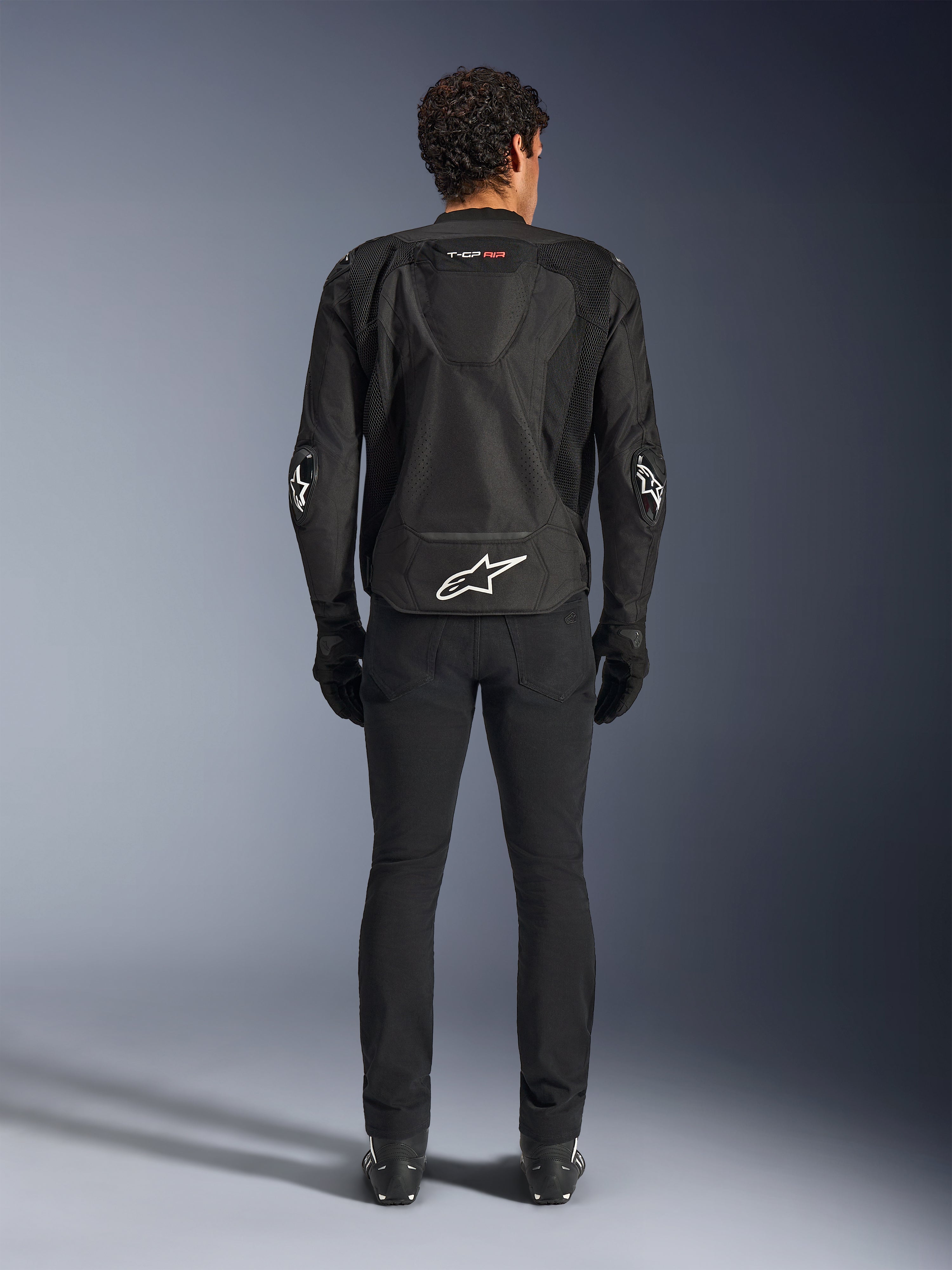 Alpinestars T-GP Air Jacket, zwart, achteraanzicht van een man die de textiele motorjas draagt, voorzien van mesh ventilatiepanelen, een aerodynamische bult en witte Alpinestars-logo's op de onderrug en ellebogen, gedragen met een zwarte motorbroek en handschoenen
