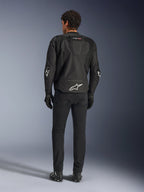Alpinestars T-GP Air Jacket, zwarte motorjas, achteraanzicht op model, toont 3D mesh ventilatiepanelen en aerodynamische bult, voorzien van wit sterlogo op de onderrug en ellebogen, gedragen met zwarte motorbroek en handschoenen, sportieve rijkleding.
