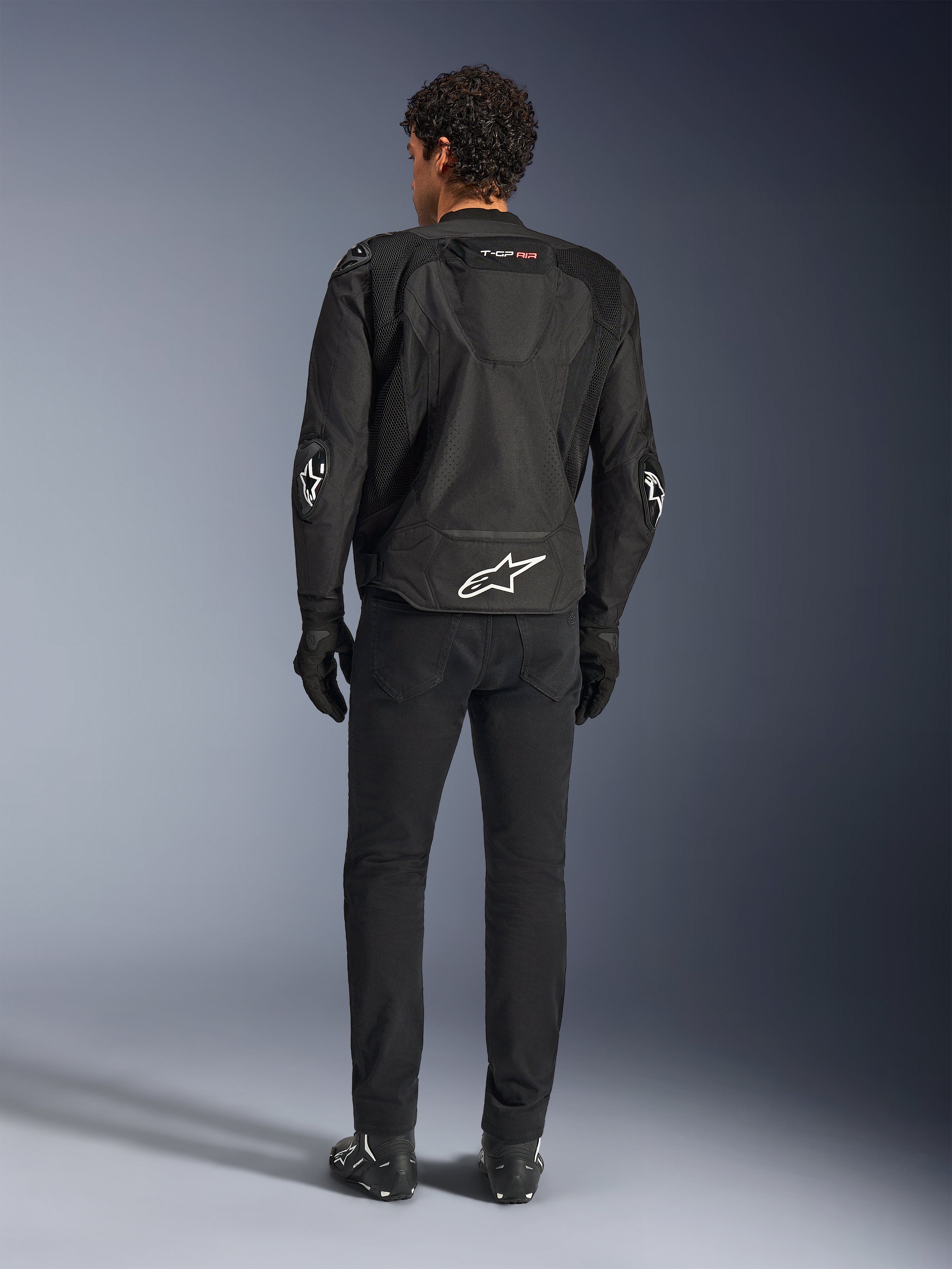Alpinestars T-GP Air Jacket, zwarte motorjas, achteraanzicht op model, toont 3D mesh ventilatiepanelen en aerodynamische bult, voorzien van wit sterlogo op de onderrug en ellebogen, gedragen met zwarte motorbroek en handschoenen, sportieve rijkleding.