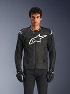 Alpinestars T-GP Air Jacket, zwarte textielen motorjas met een groot wit sterlogo op de borst, voorzien van geperforeerde ventilatiepanelen en 3D-mesh, externe schoudersliders, gedragen met zwart-witte handschoenen, strak sportief ontwerp voor rijden op de weg bij warm weer