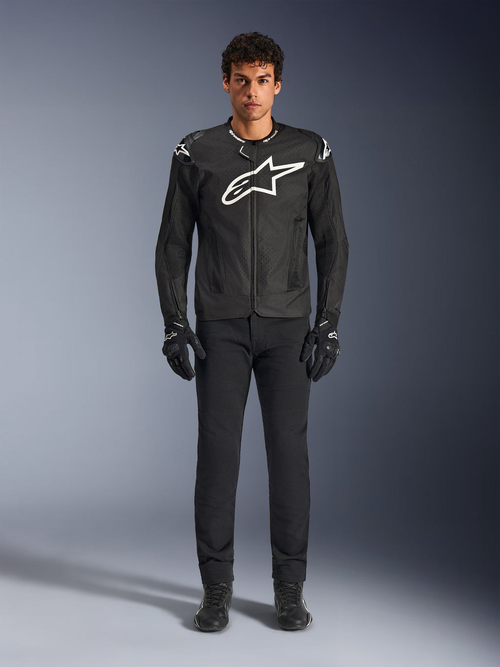 Alpinestars T-GP Air Jacket, zwarte textielen motorjas met een groot wit Astars-logo op de borst, gedragen door een model met zwarte motorbroek en handschoenen, voorzien van geperforeerde panelen en mesh-ventilatie voor sportief rijden